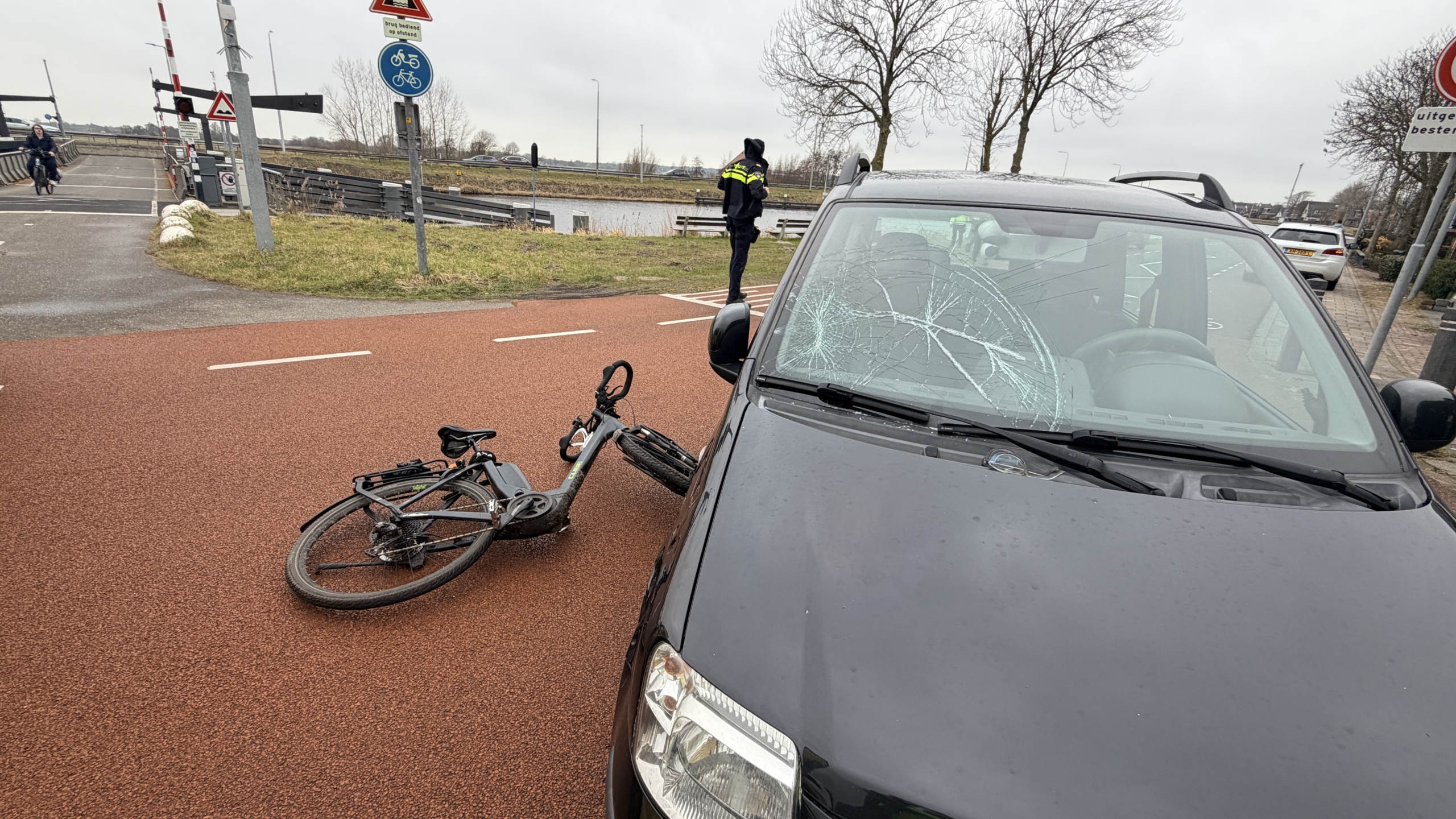 Auto met gebarsten voorruit en liggende fiets op fietspad, agent op de achtergrond.