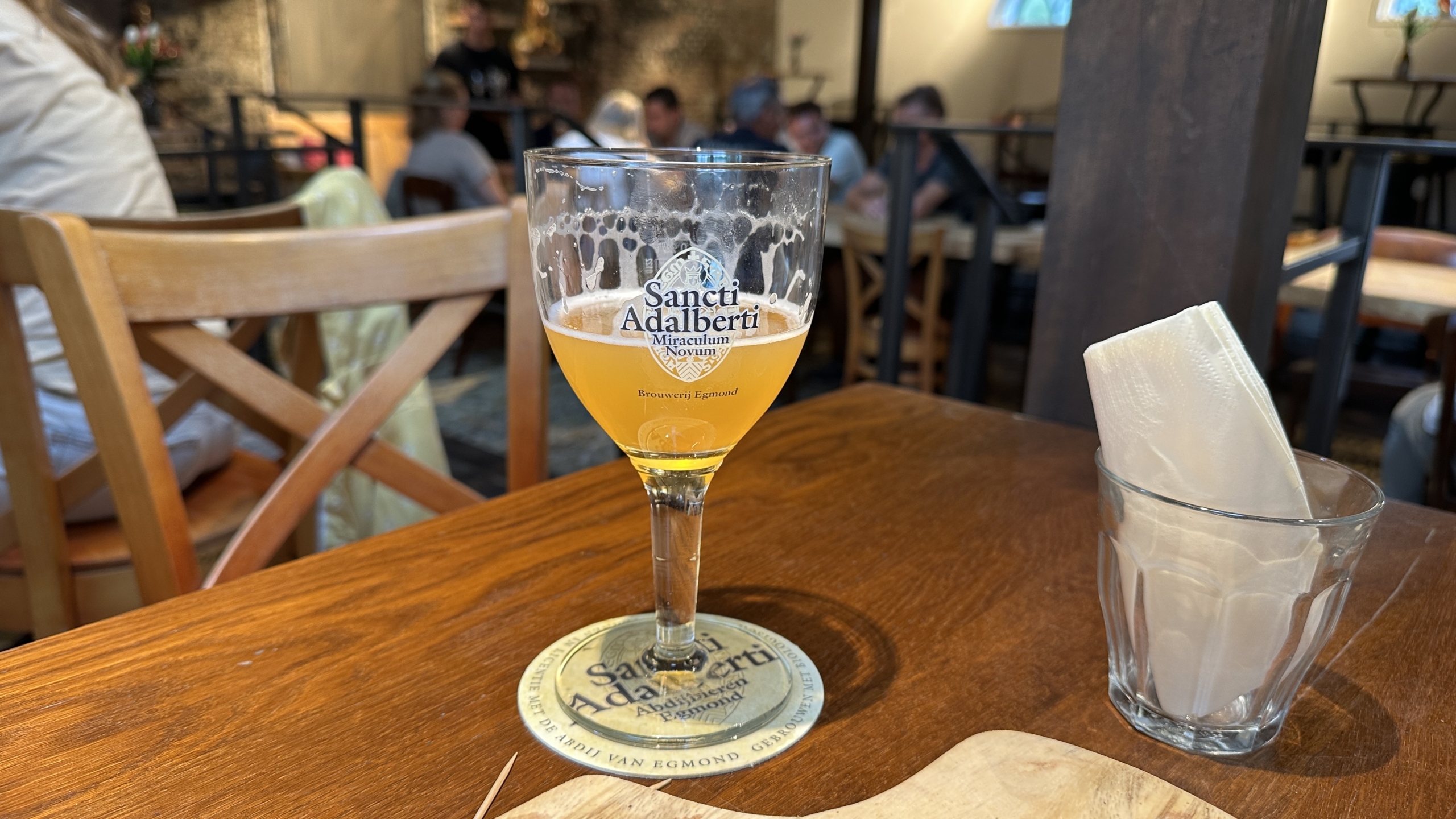 Glas met oranje bier van Sancti Adalberti op een houten tafel in een café, met mensen op de achtergrond.