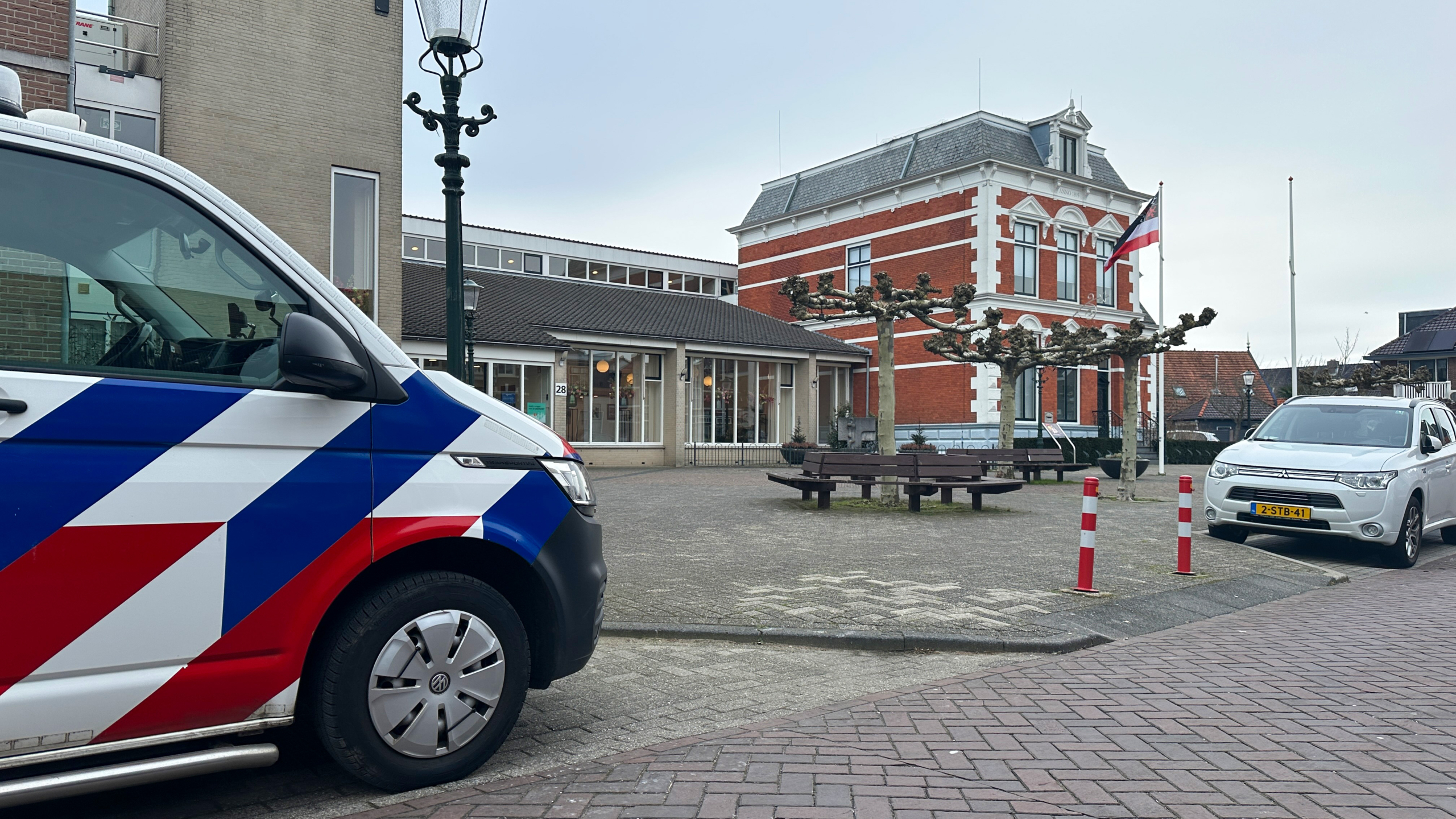 Een politiebus met rood, wit en blauwe strepen staat geparkeerd bij een bakstenen gebouw met een Nederlandse vlag.