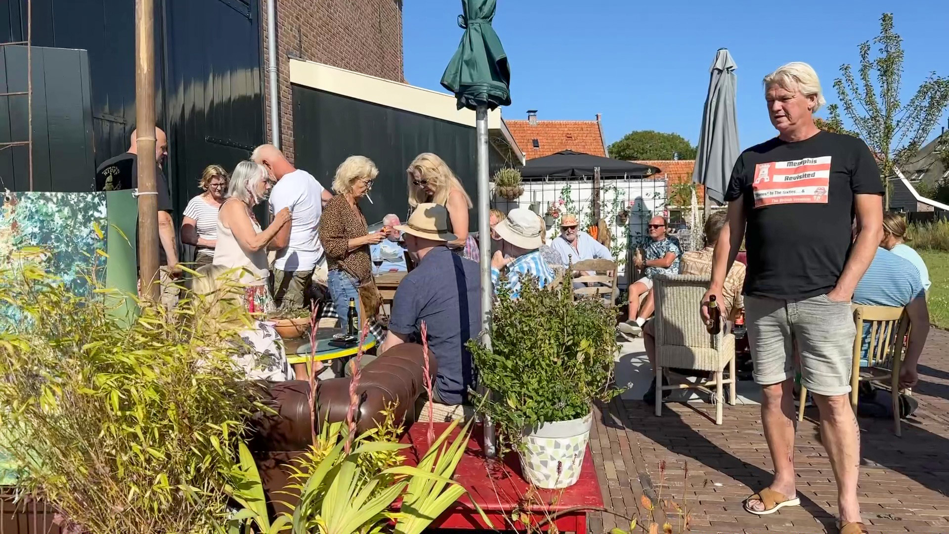 Mensen genieten van een zonnige dag op een druk terras, omgeven door planten en met tafels en stoelen.