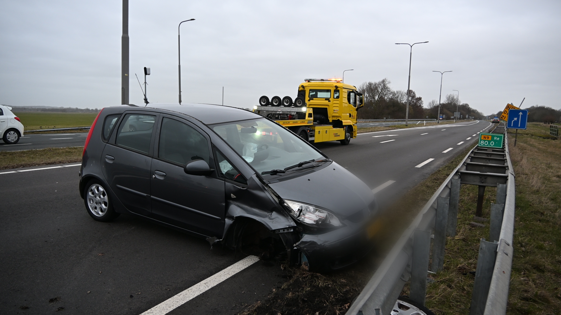 Auto met schade aan de voorzijde naast een vangrail op de snelweg, met een gele sleepwagen op de achtergrond.