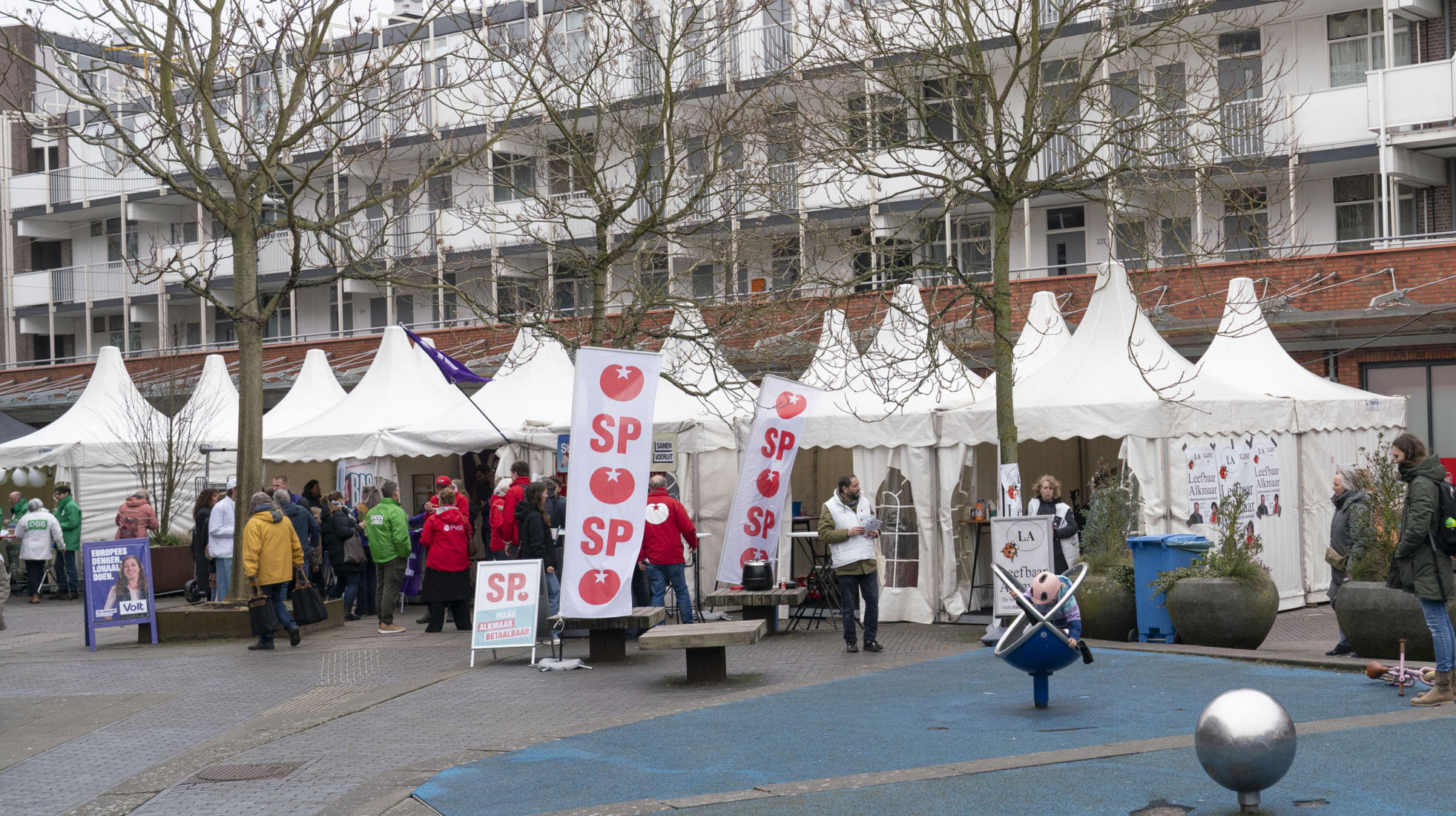 Straatbeeld met partijtenten en SP-promotiemateriaal, omgeven door mensen en enkele bomen.