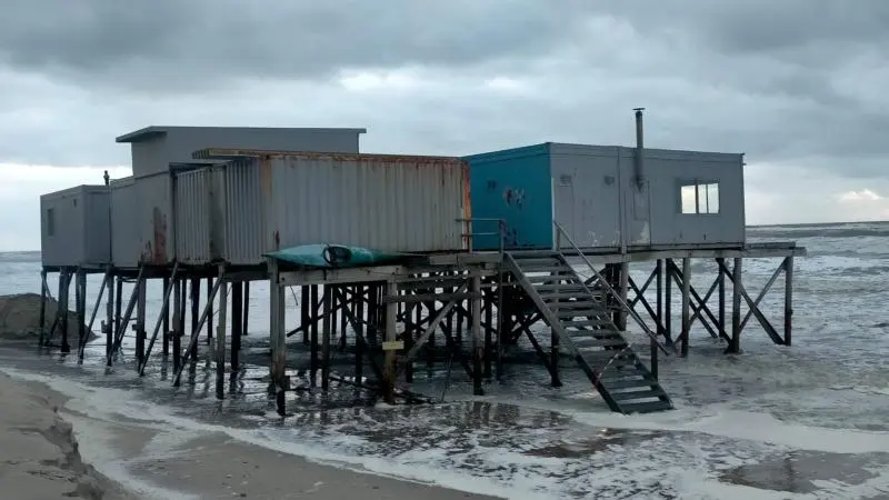 Verhoogde gebouwen op palen aan zee met bewolkte lucht op de achtergrond.