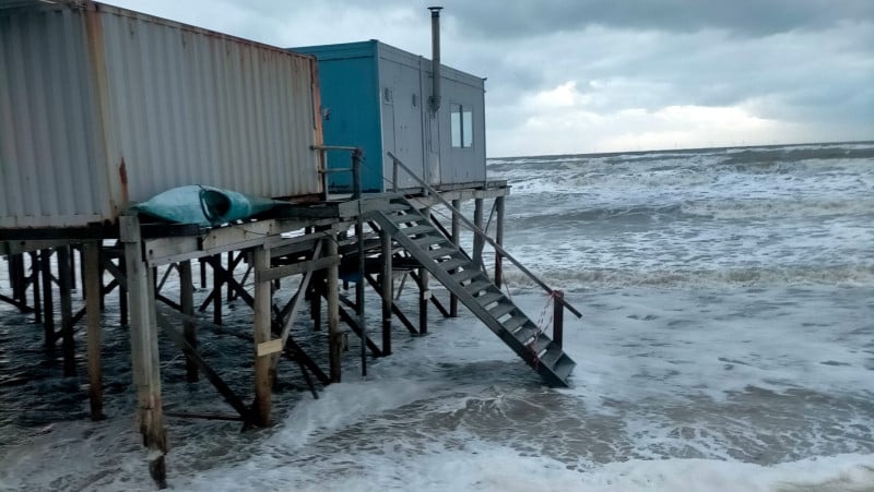 Houten constructie op palen boven de zee, met golven die tegen de onderkant slaan en een bewolkte hemel op de achtergrond.