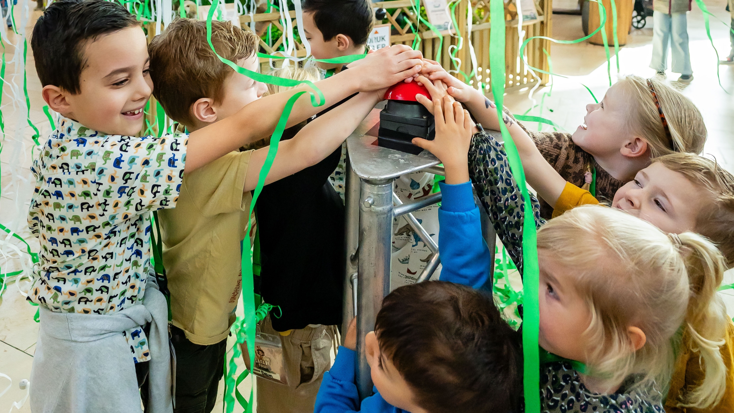 Een groep kinderen drukt samen op een rode knop, omgeven door groene en witte slingers.