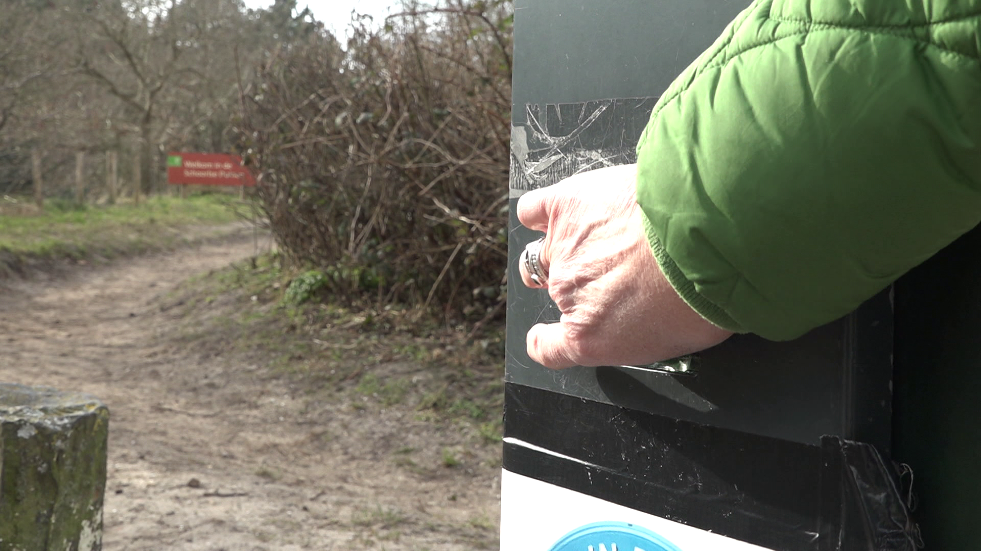 Een hand in een groene jas opent een groene kast naast een onverharde weg met een wazig bord op de achtergrond.
