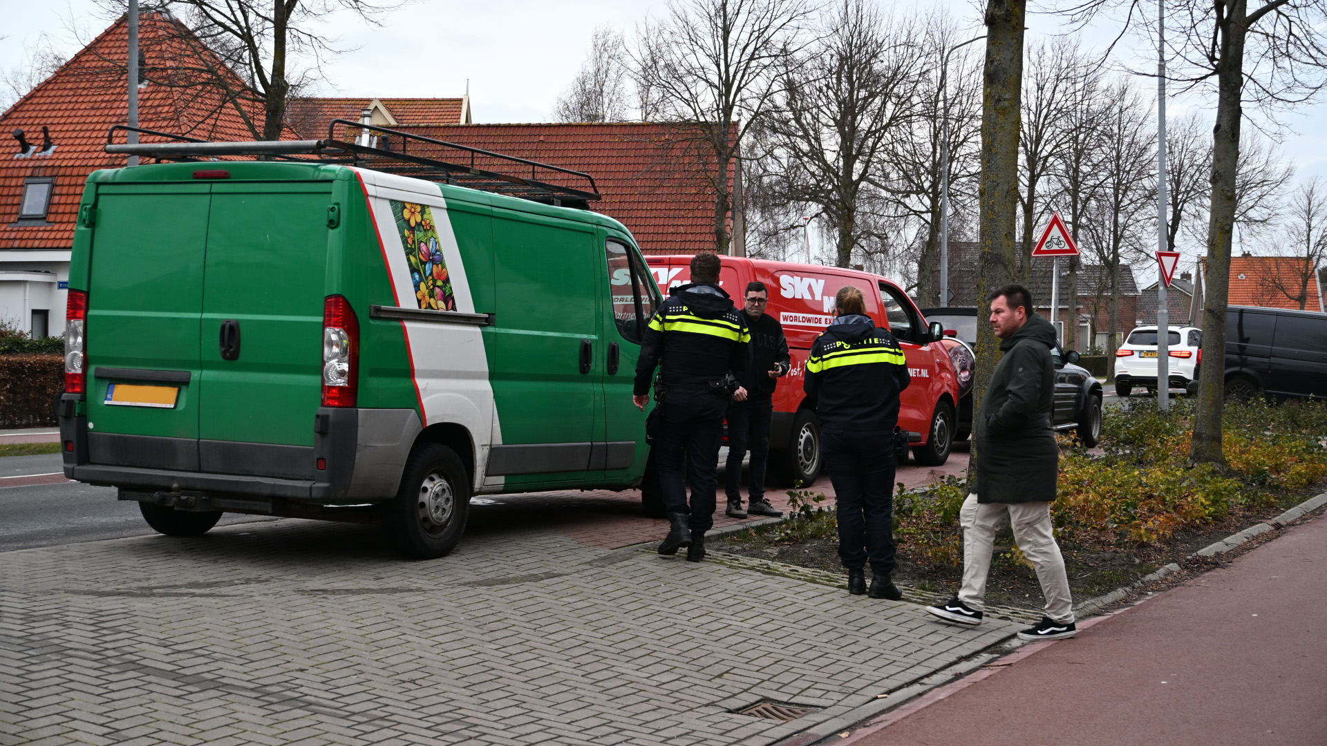 Politieagenten staan bij een groene en een rode bestelwagen naast een trottoir, terwijl een man toekijkt.