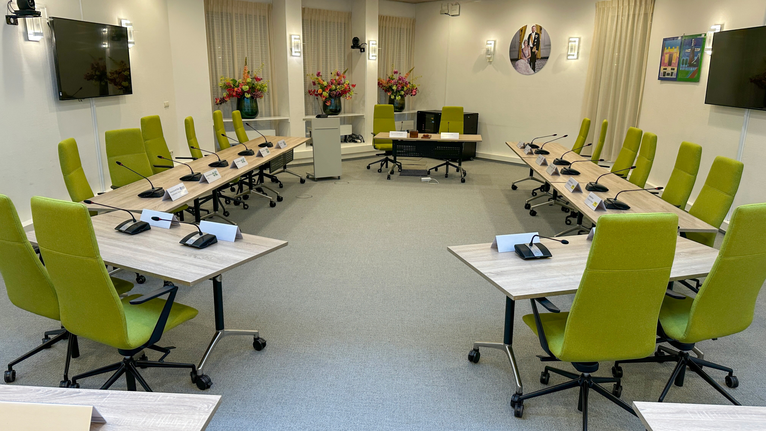 Vergaderruimte met groene stoelen, microfoons op tafels in U-vorm, bloemen op de achtergrond en foto aan de muur.