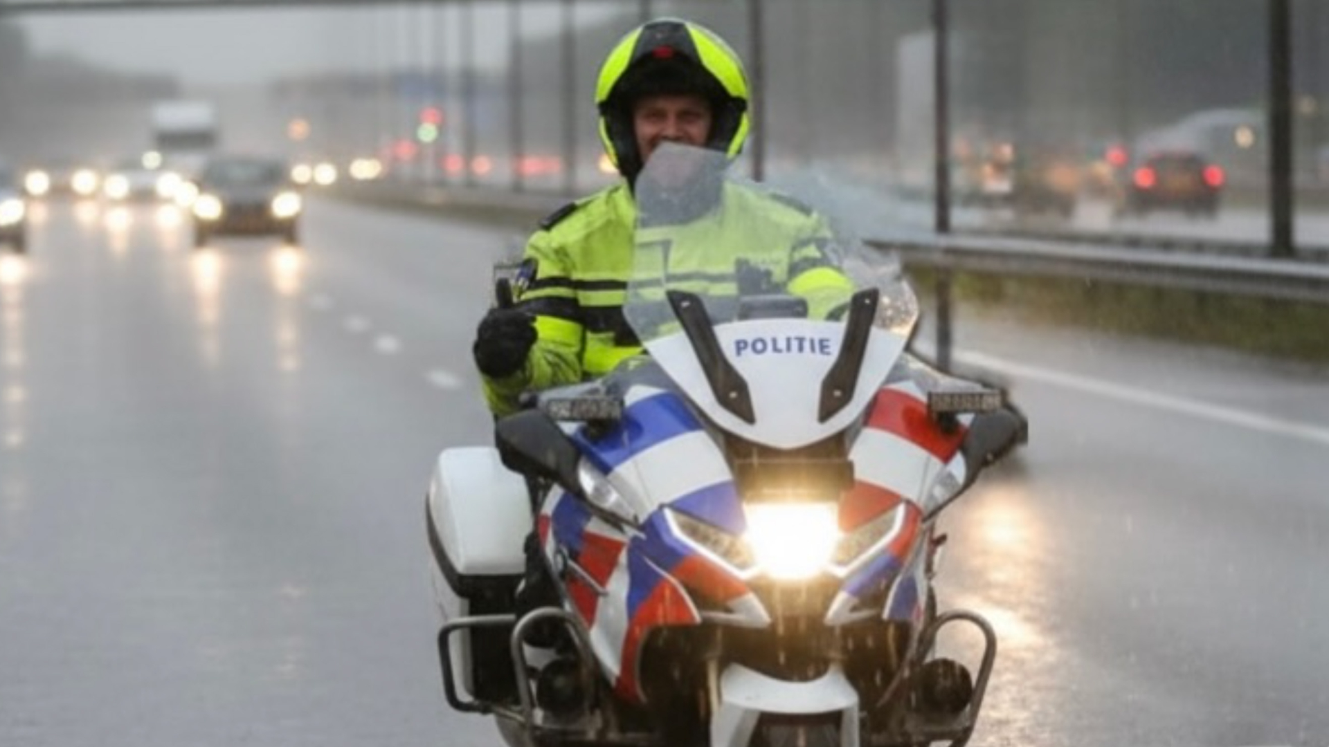 Politiemotoragent met helm in regen op snelweg, op herkenbare motor met rood-blauwe strepen.