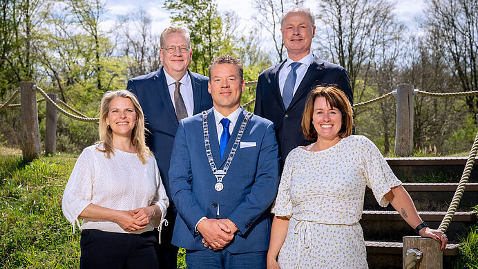 Vijf mensen poseren in formele kleding buiten op een trap met bomen op de achtergrond.