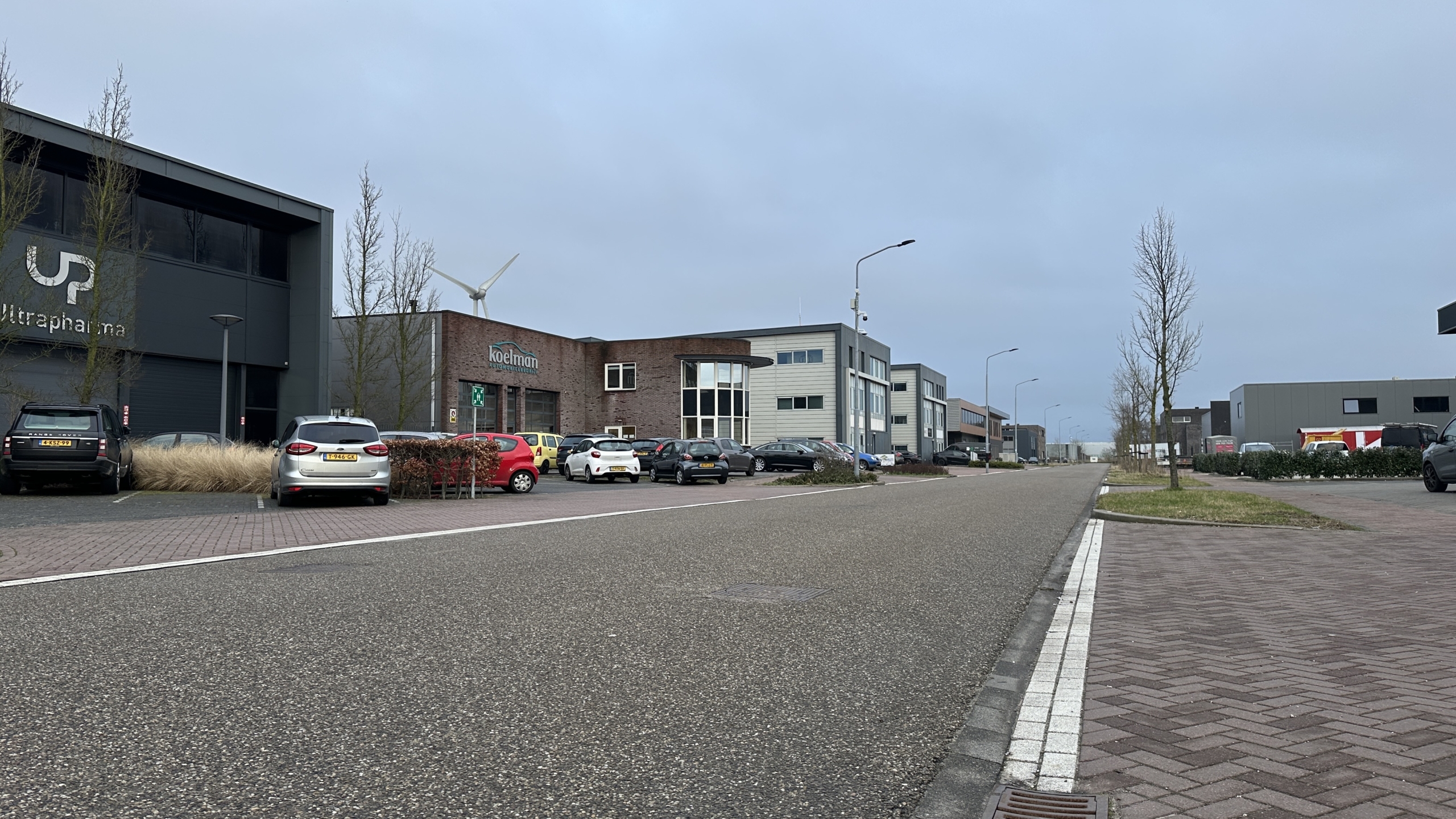 Industriegebied met bedrijfspanden en geparkeerde auto's langs een weg, onder een bewolkte hemel.