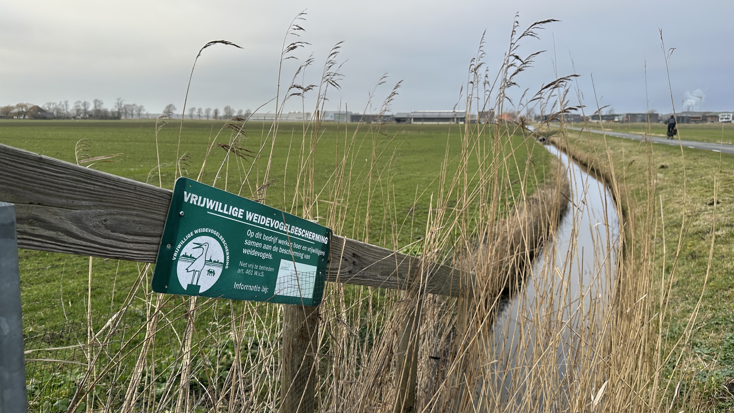 Bordje met "Vrijwillige Weidevogelbescherming" bij een groen weiland met sloot en fietser op de achtergrond.