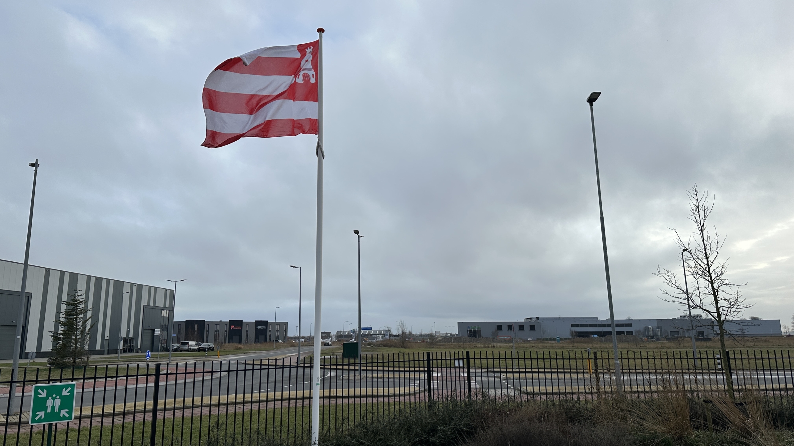 Een rood-wit gestreepte vlag wappert in de wind voor industriële gebouwen onder een bewolkte hemel.
