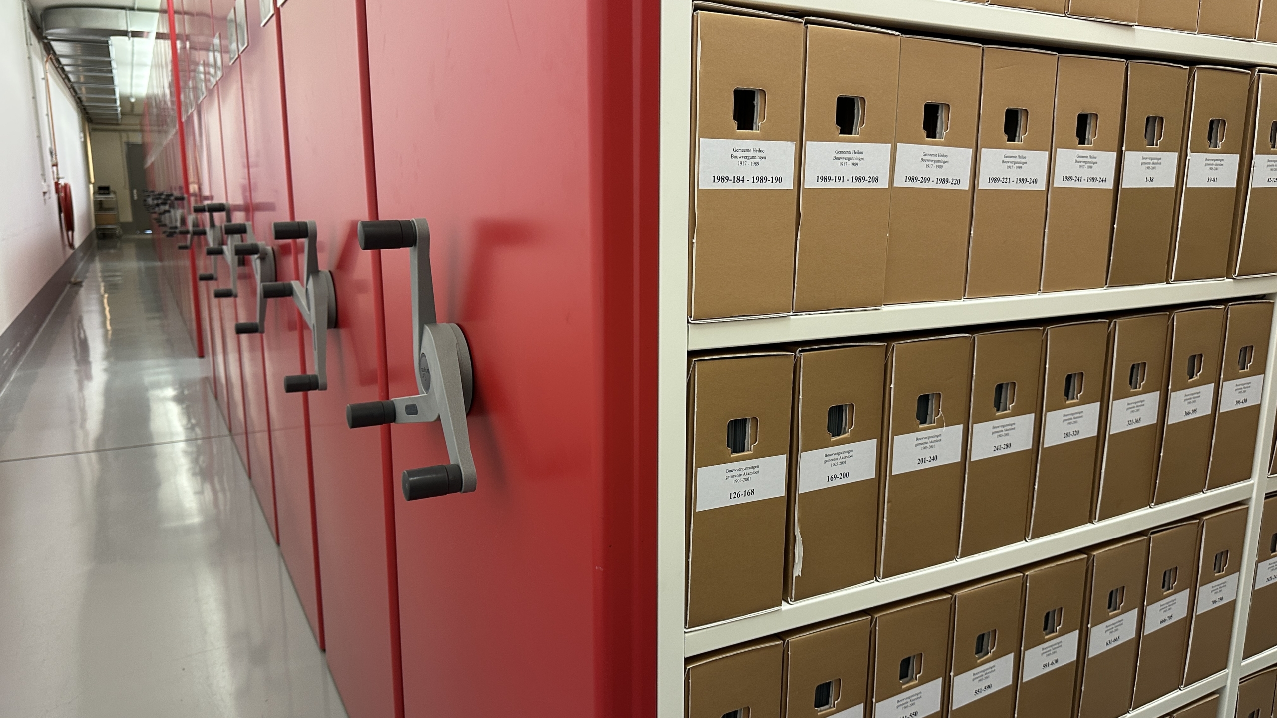 Rijen archiefkasten met bruine dozen en rode schuifmechanismen in een verlichte gang.