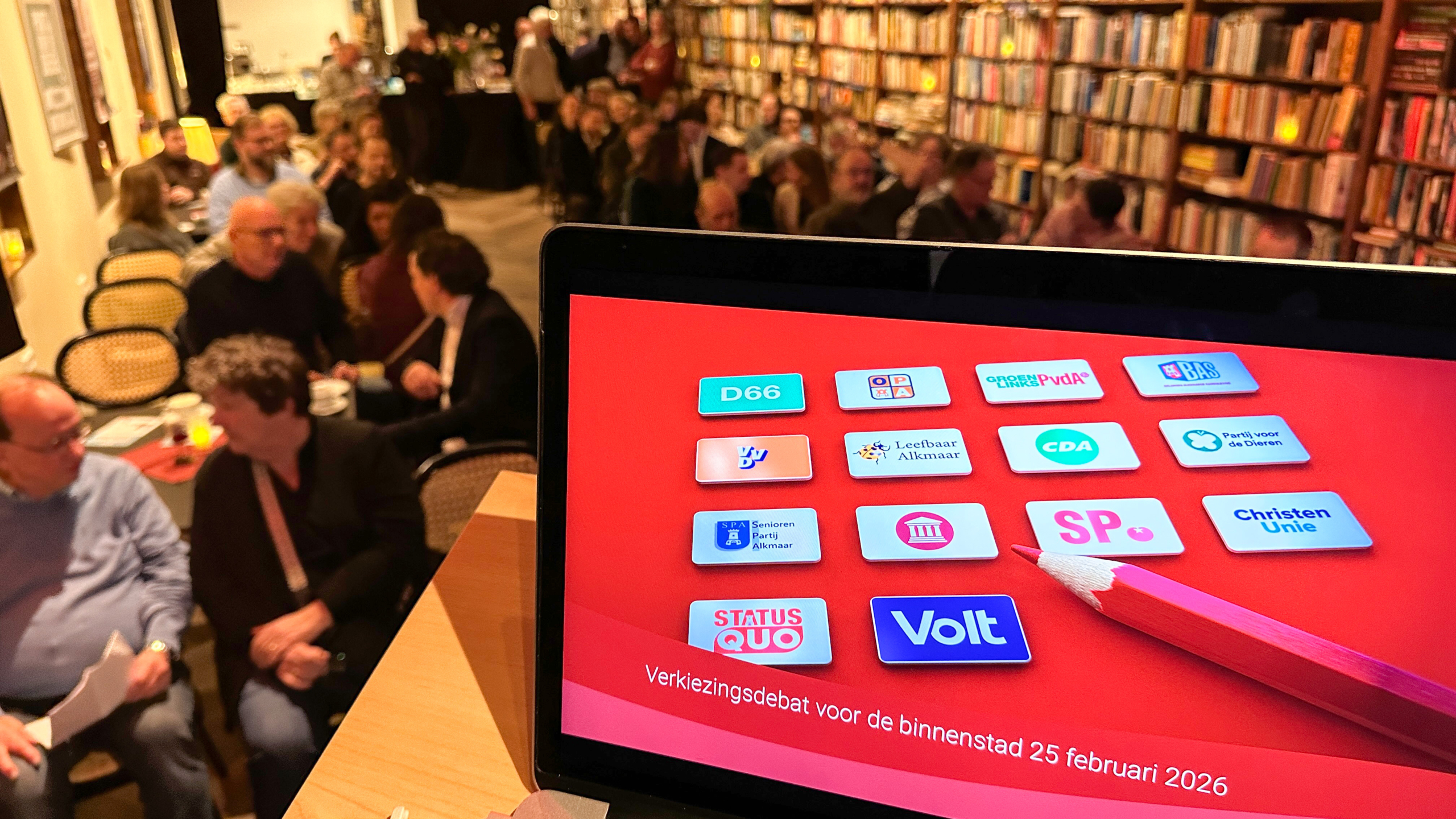 Scherm toont politieke logo's en tekst "Verkiezingsdebat voor de binnenstad 25 februari 2026" in een ruimte met mensen.