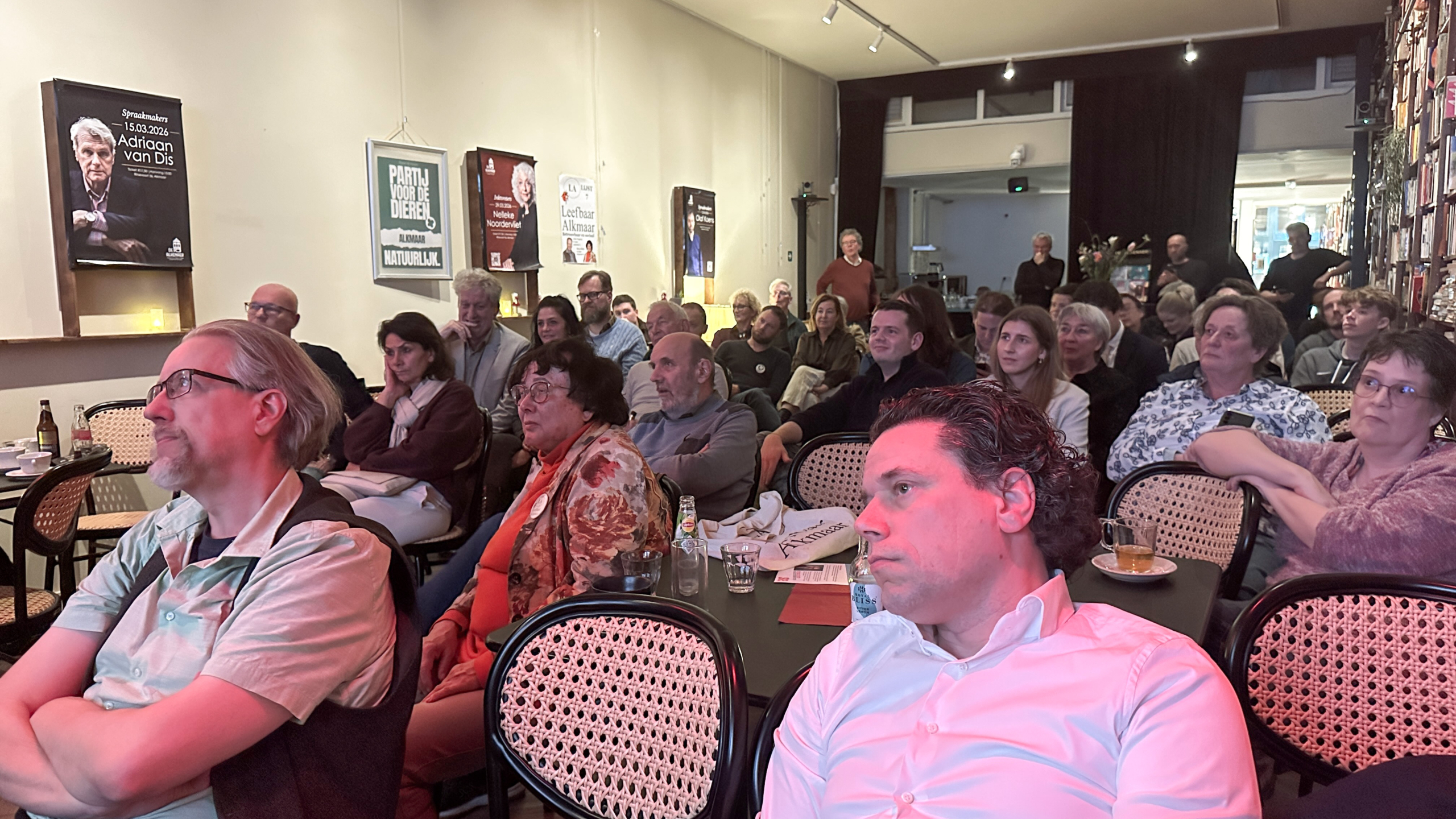 Een groep mensen zit aandachtig te luisteren tijdens een presentatie of bijeenkomst in een zaal met posters aan de muur.