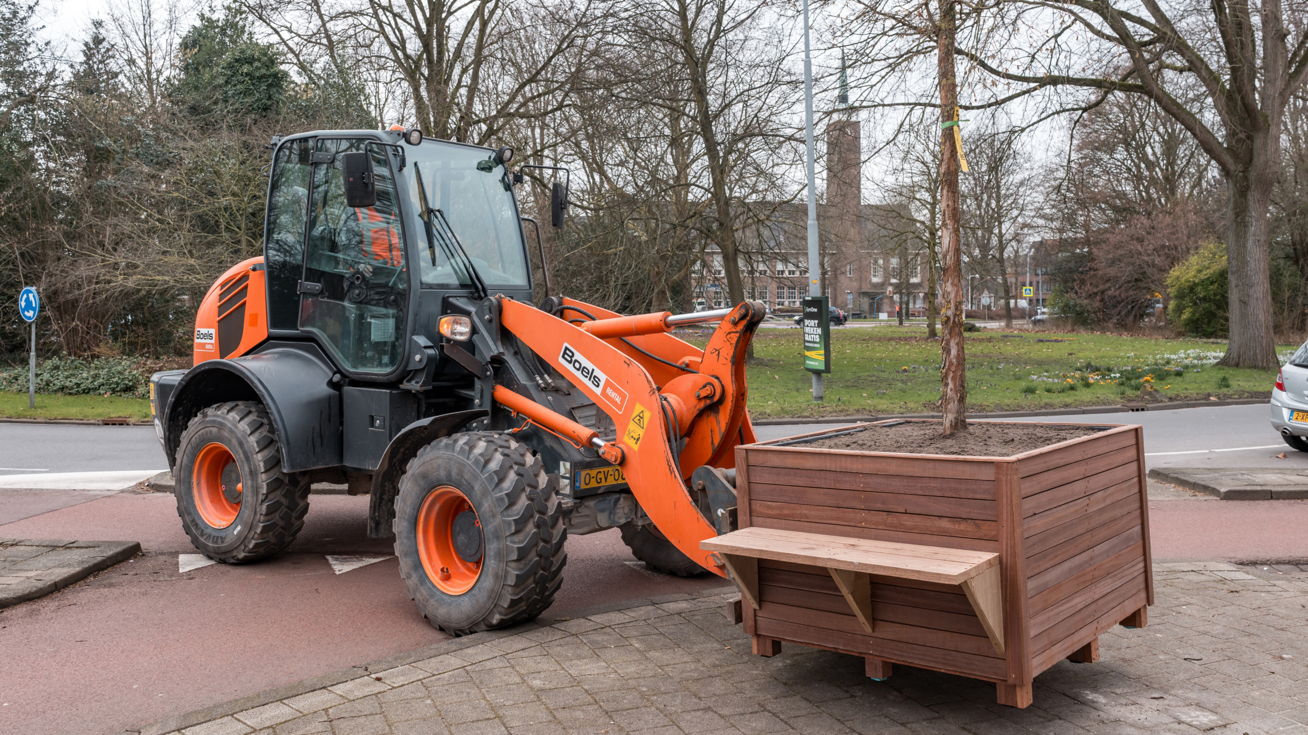 Graafmachine van Boels plaatst een houten plantenbak met boom op een trottoir nabij een rotonde.