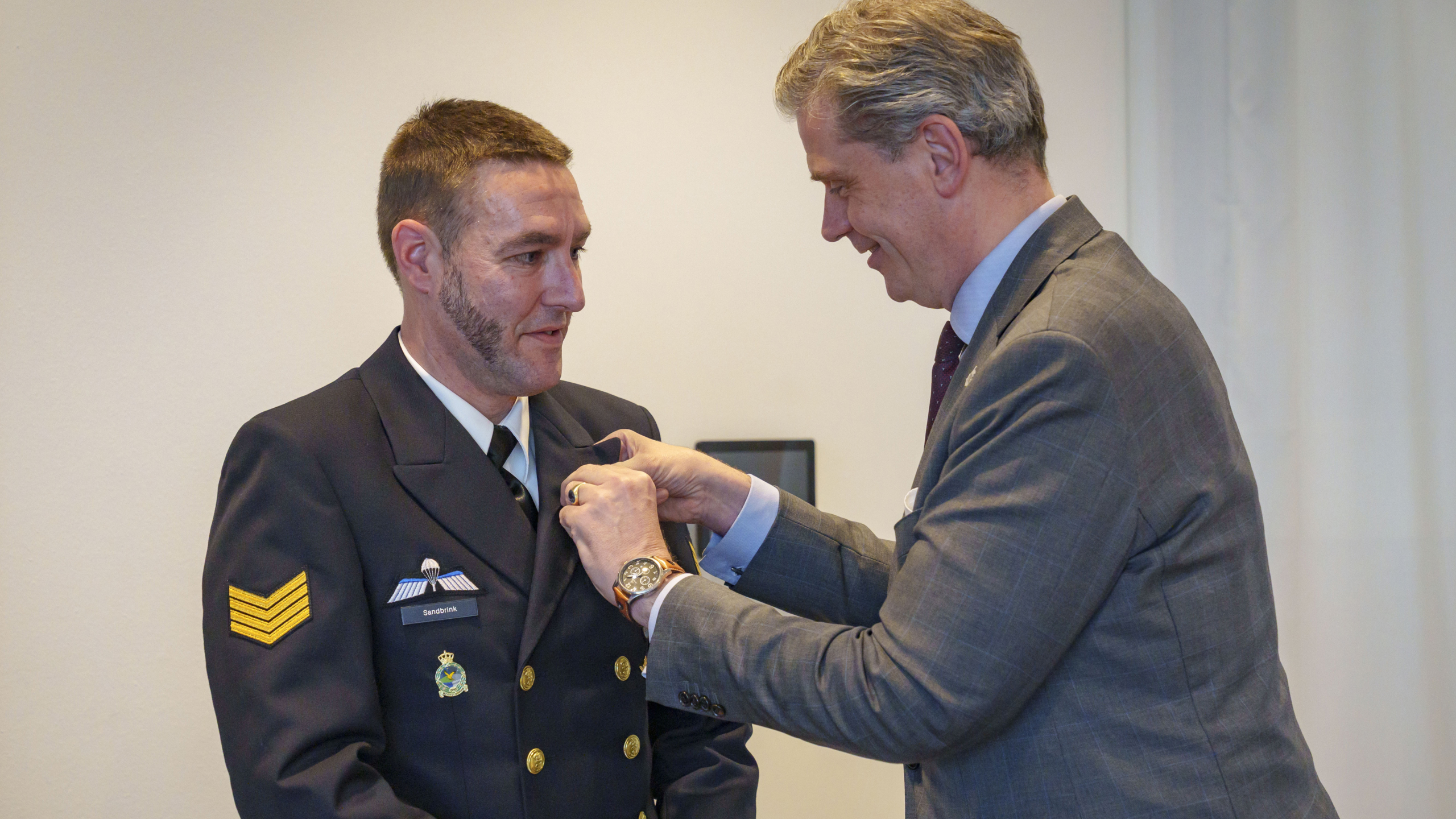 Een man in uniform ontvangt een onderscheiding van een andere man in pak in een formele setting.