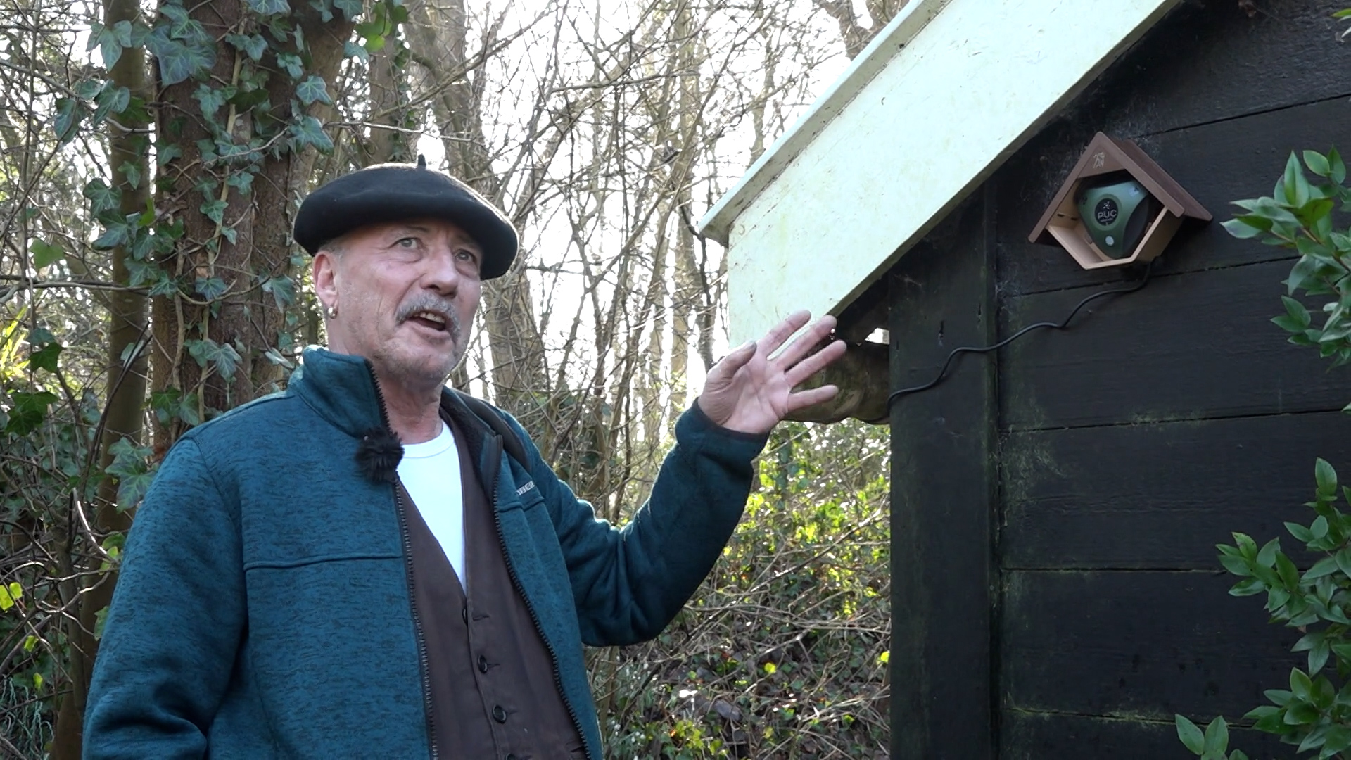 Man met baret wijst naar een vogelkastje met camera aan een houten schuur in een bosrijke omgeving.