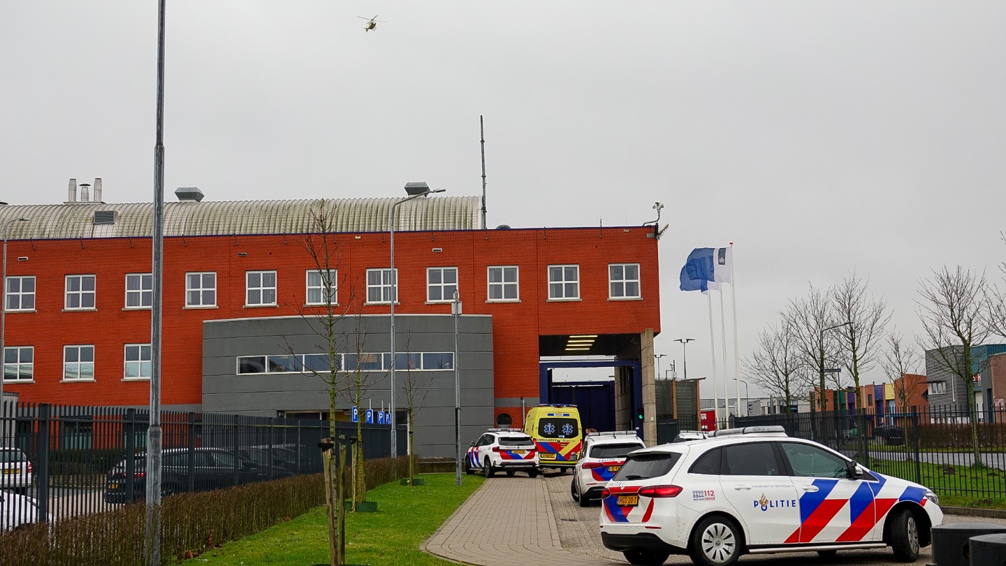Politiewagens en een ambulance bij een rood bakstenen gebouw met vlaggen en een helikopter in de lucht.