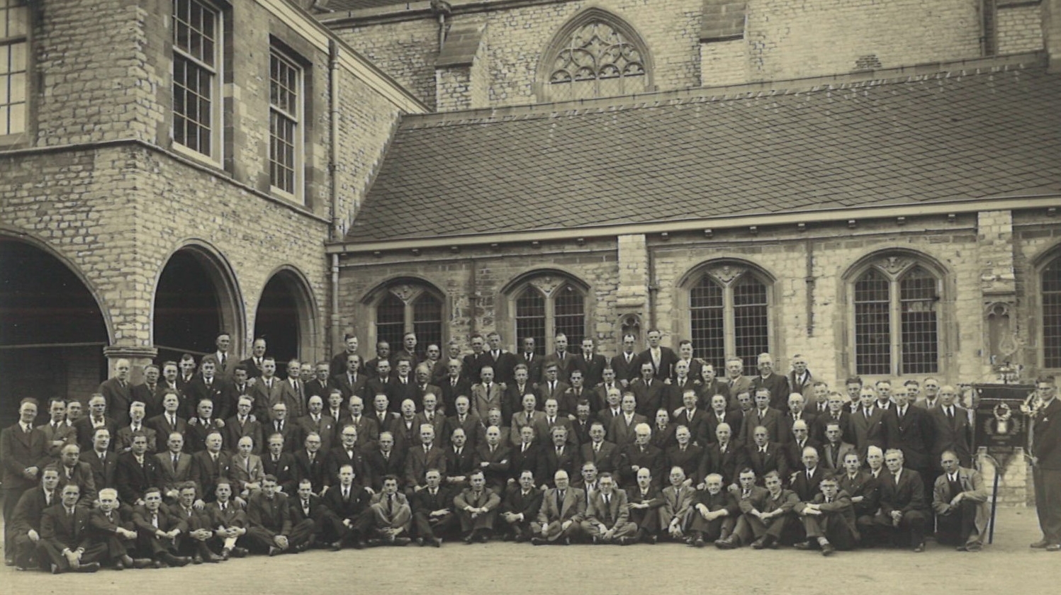 Een grote groep mannen poseert voor een historisch gebouw met ramen in gotische stijl.