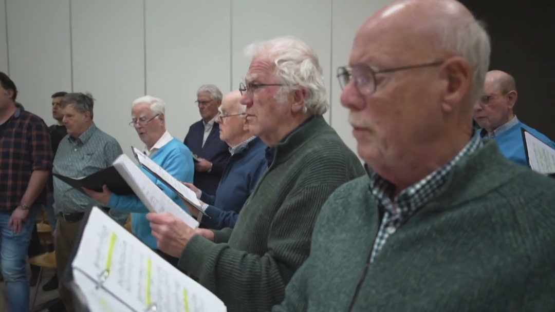Mannengroep zingt samen in een repetitielokaal, elk met muziekbladen in de hand.