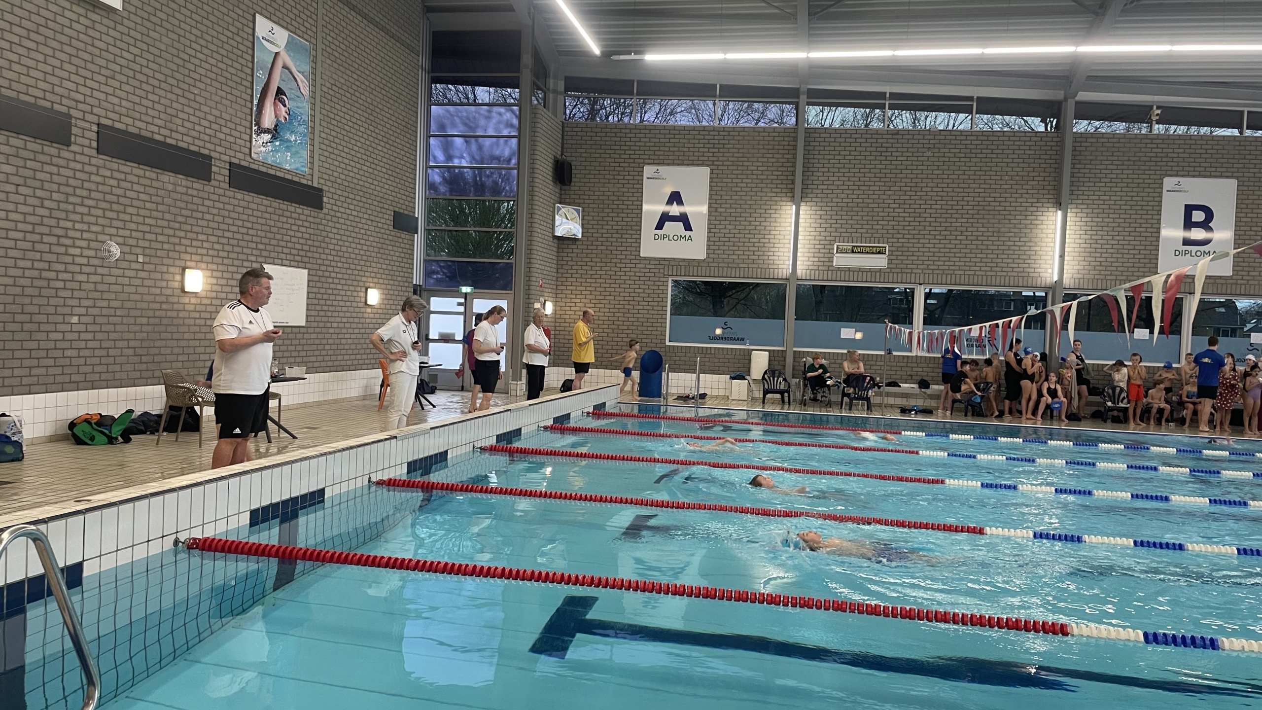 Zwemwedstrijd in een overdekt zwembad met juryleden langs de kant en toeschouwers op de achtergrond.