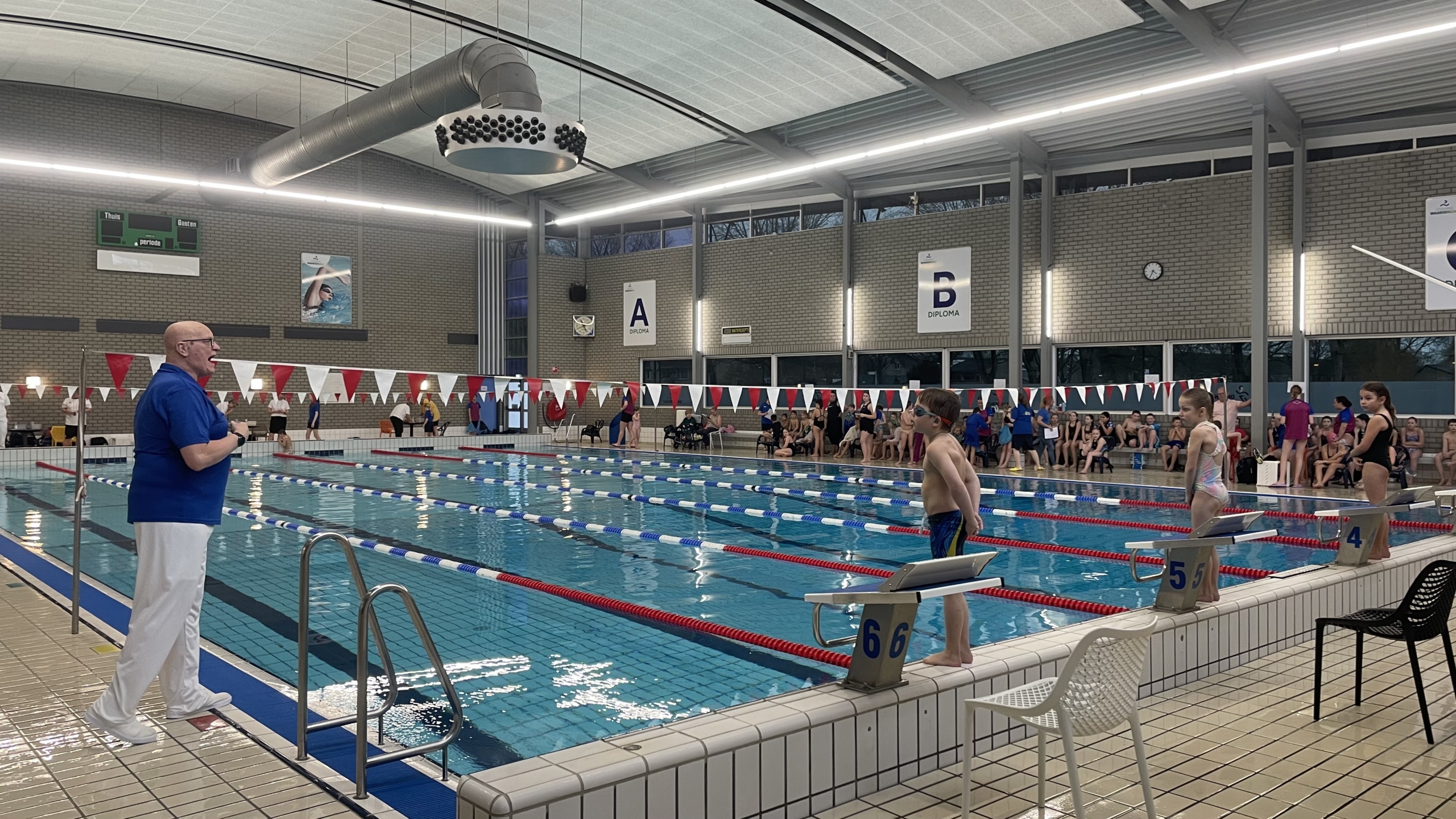 Kinderen staan klaar voor een zwemwedstrijd in een overdekt zwembad, terwijl een official bij het startblok staat.