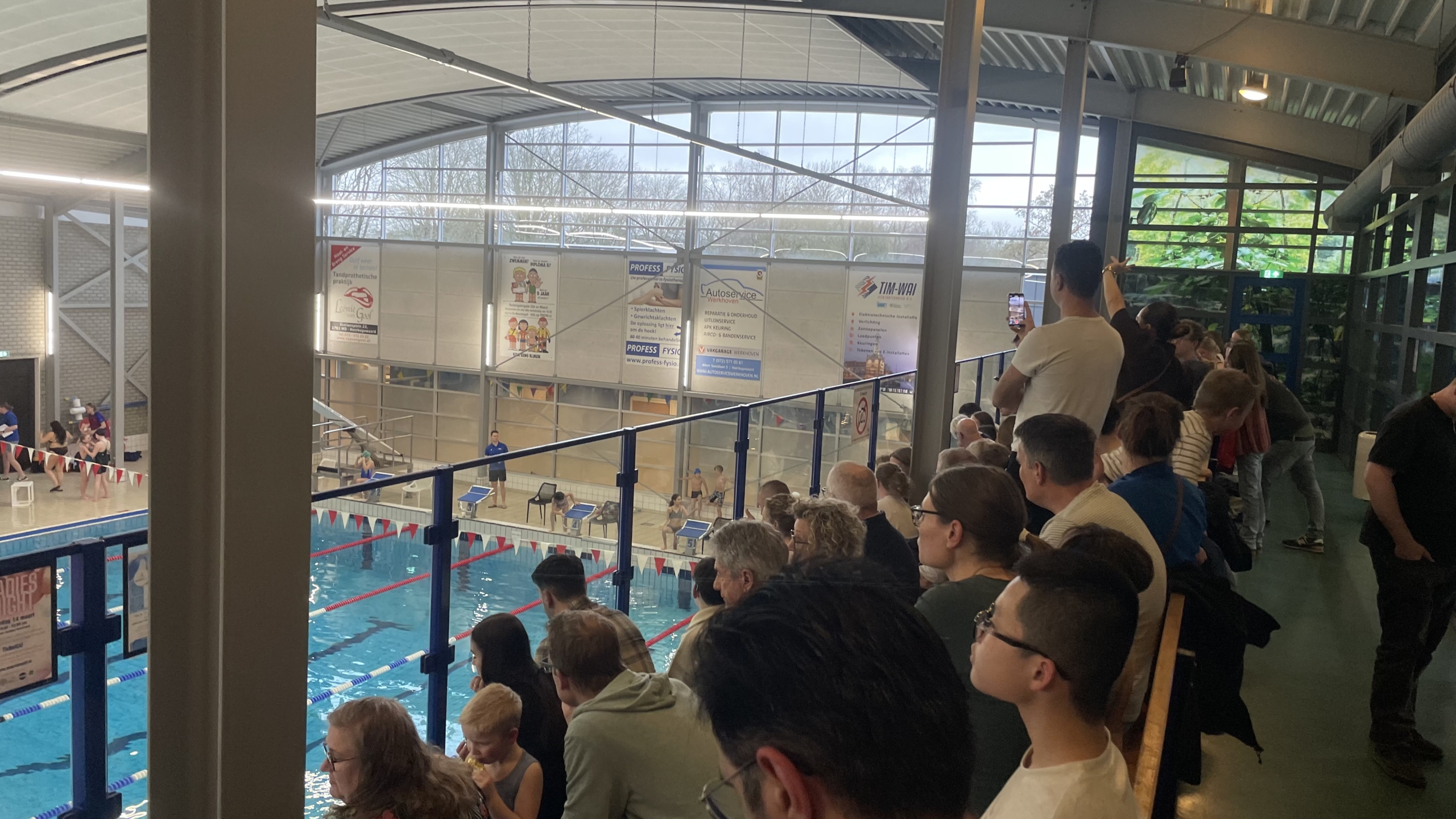 Toeschouwers zitten op tribunes bij een zwembad, waar een zwemwedstrijd lijkt te plaatsvinden.