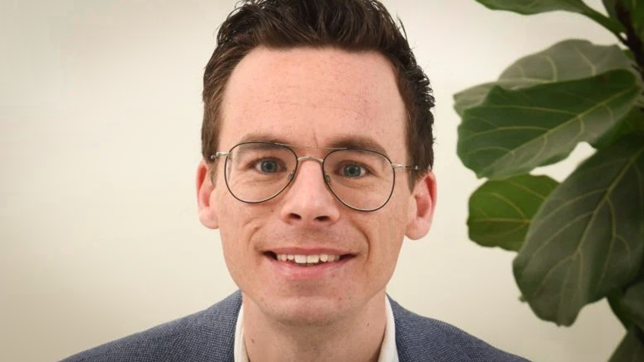 Man met bril en donker haar glimlacht naar de camera, met een plant op de achtergrond.
