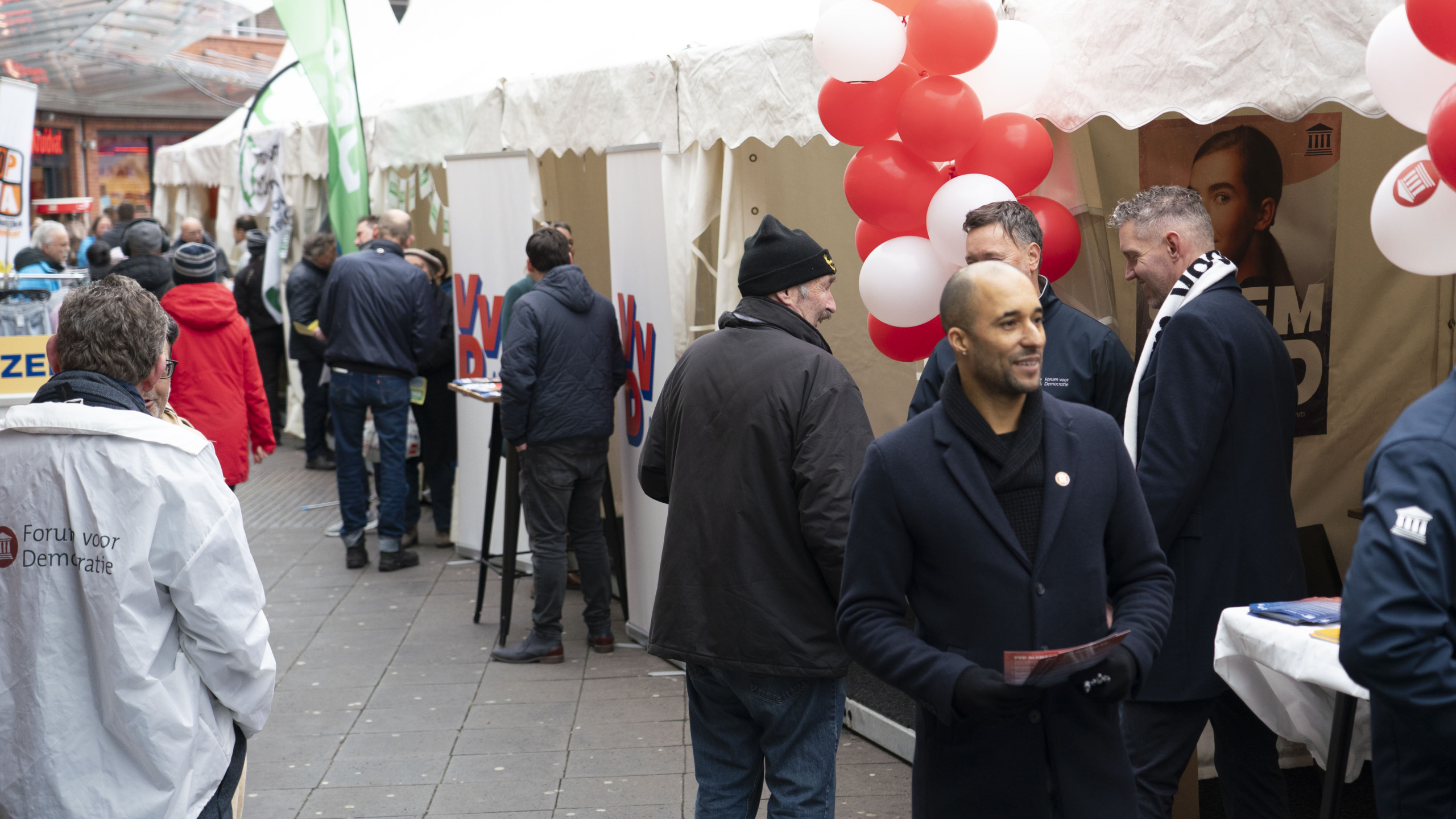 Mensen bij een politieke bijeenkomst met stands en spandoeken, waaronder een "Forum voor Democratie"-jas en rode en witte ballonnen.