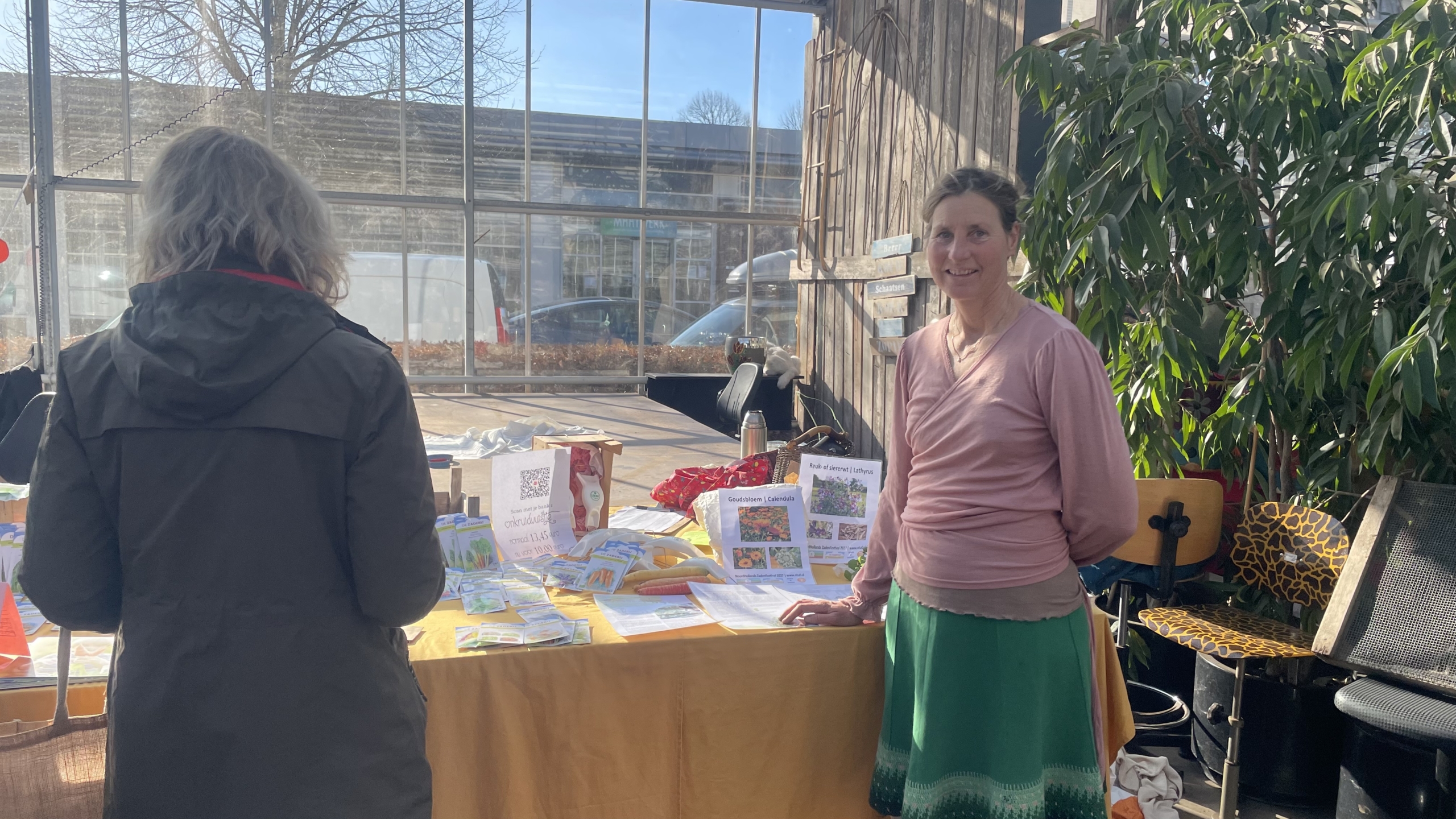 Vrouw staat bij een tafel met brochures en potten in een zonnige ruimte met grote planten en een glazen wand.