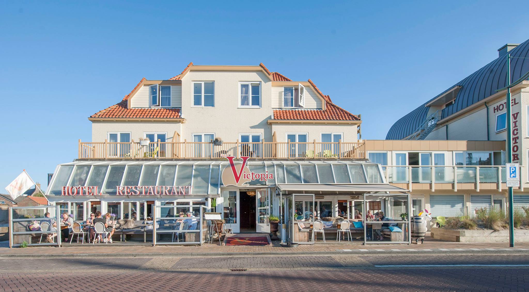 Hotel en restaurant met terracotta daken en glazen terras, waar mensen buiten zitten onder een heldere blauwe hemel.