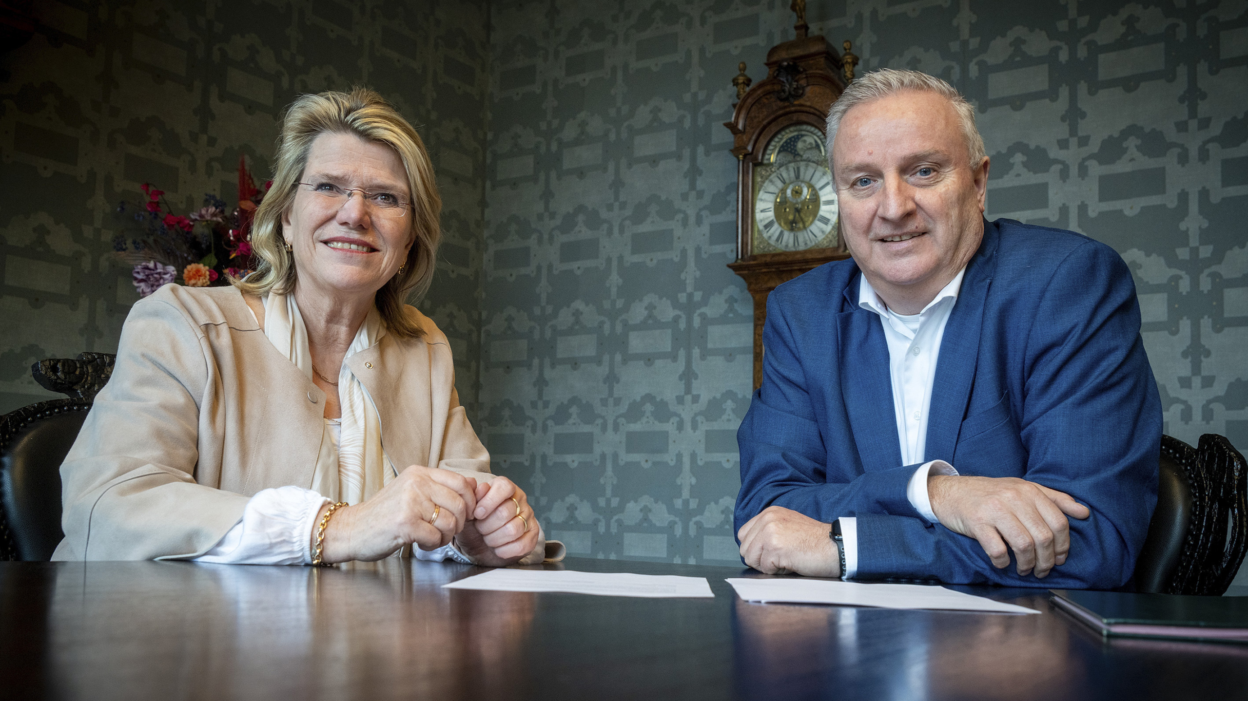 Een man en vrouw zitten aan een tafel met een klok op de achtergrond, beiden glimlachend in een formele setting.