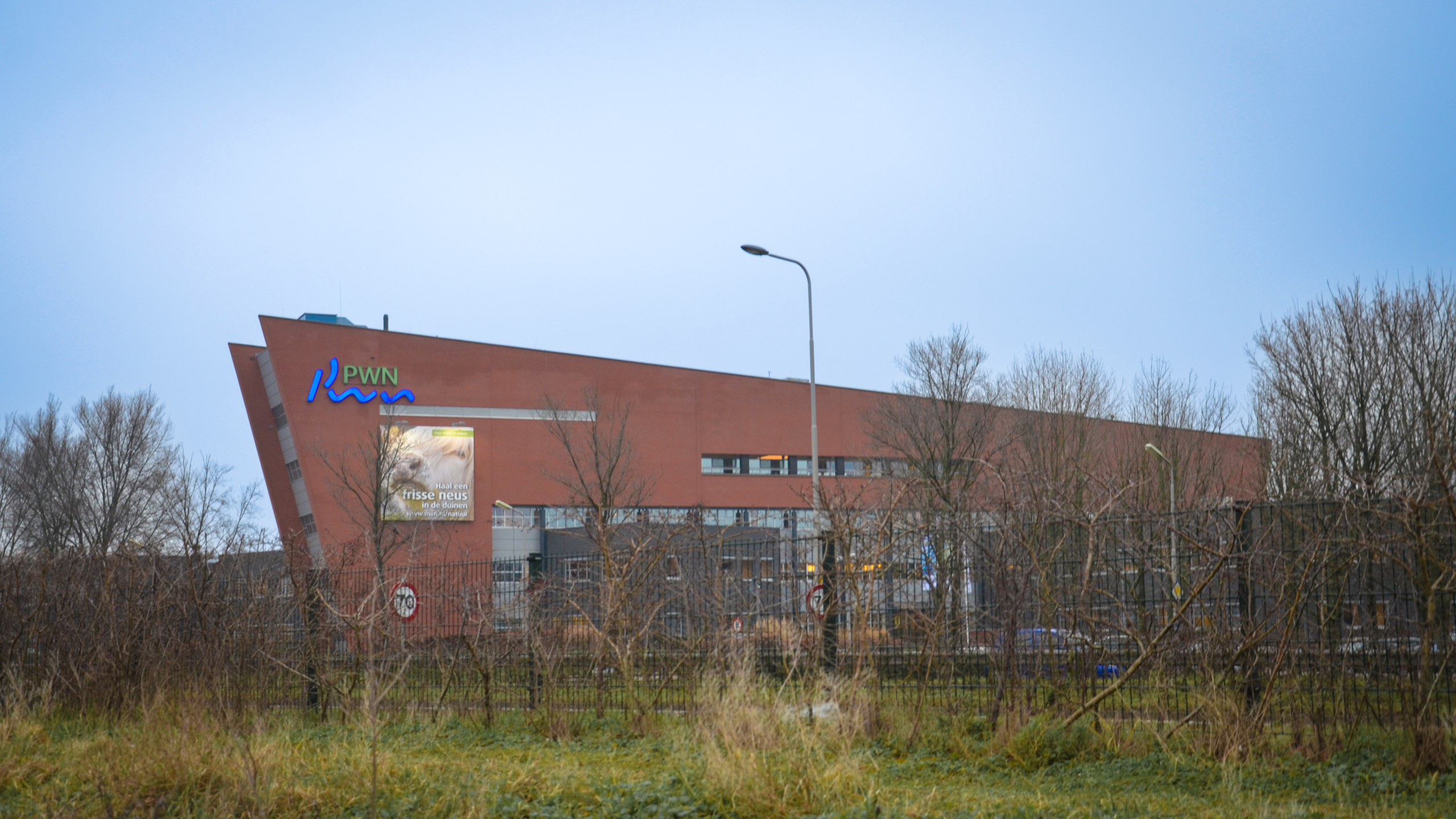 Modern gebouw met roodbruine gevel en PWN-logo, omgeven door kale bomen en grasveld.