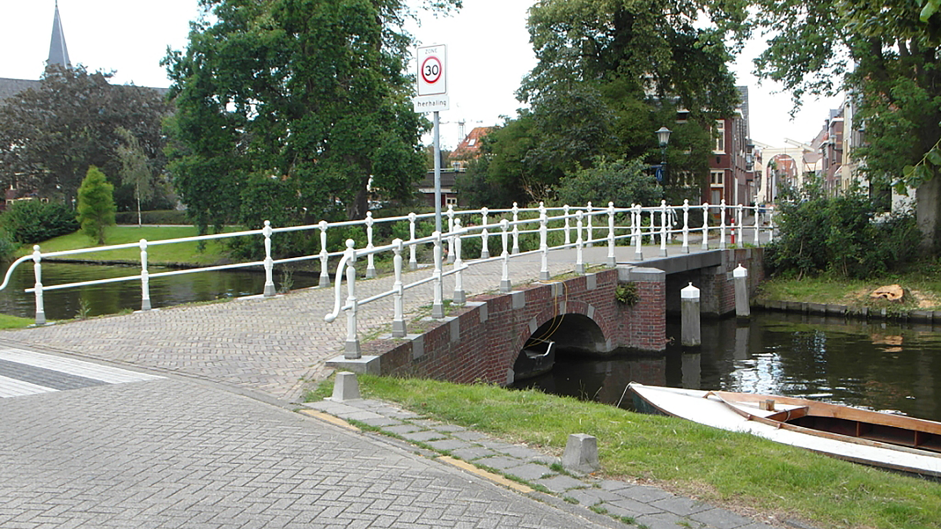 Bakstenen boogbrug over een kanaal met wit hekwerk, omringd door groen en enkele gebouwen op de achtergrond.