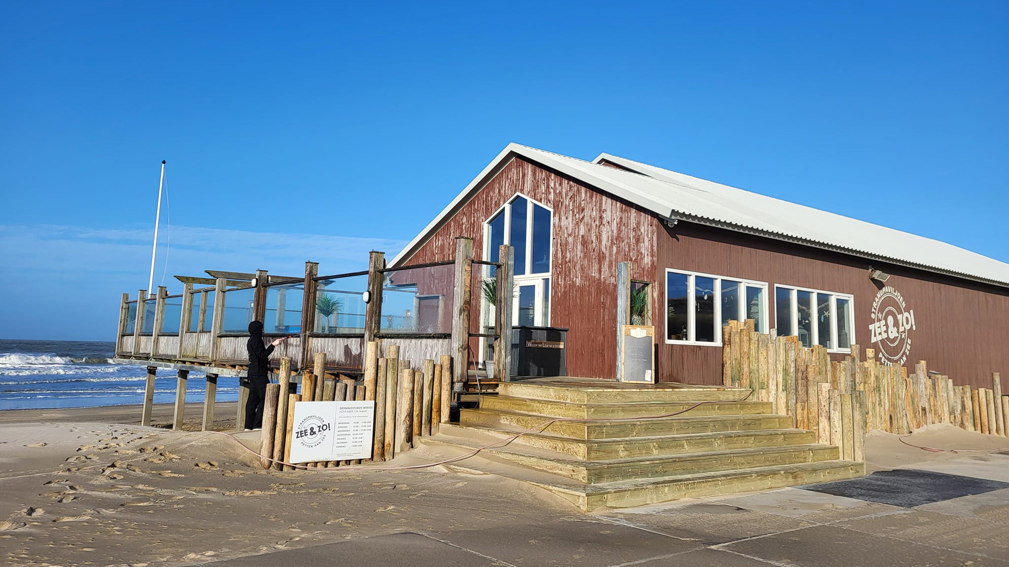 Strandpaviljoen Zee & Zo! met een houten terras, gelegen aan zee onder een blauwe hemel.