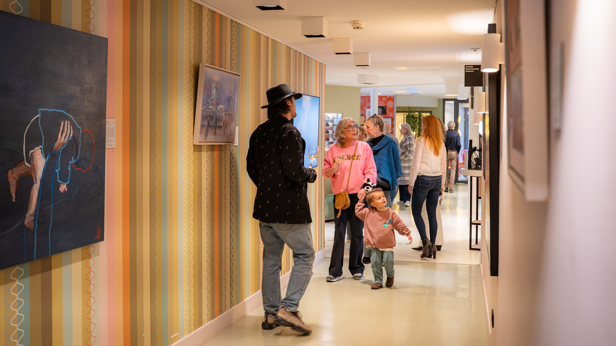 Mensen lopen door een gang met schilderijen aan de muur, een kind houdt de hand van een volwassene vast.