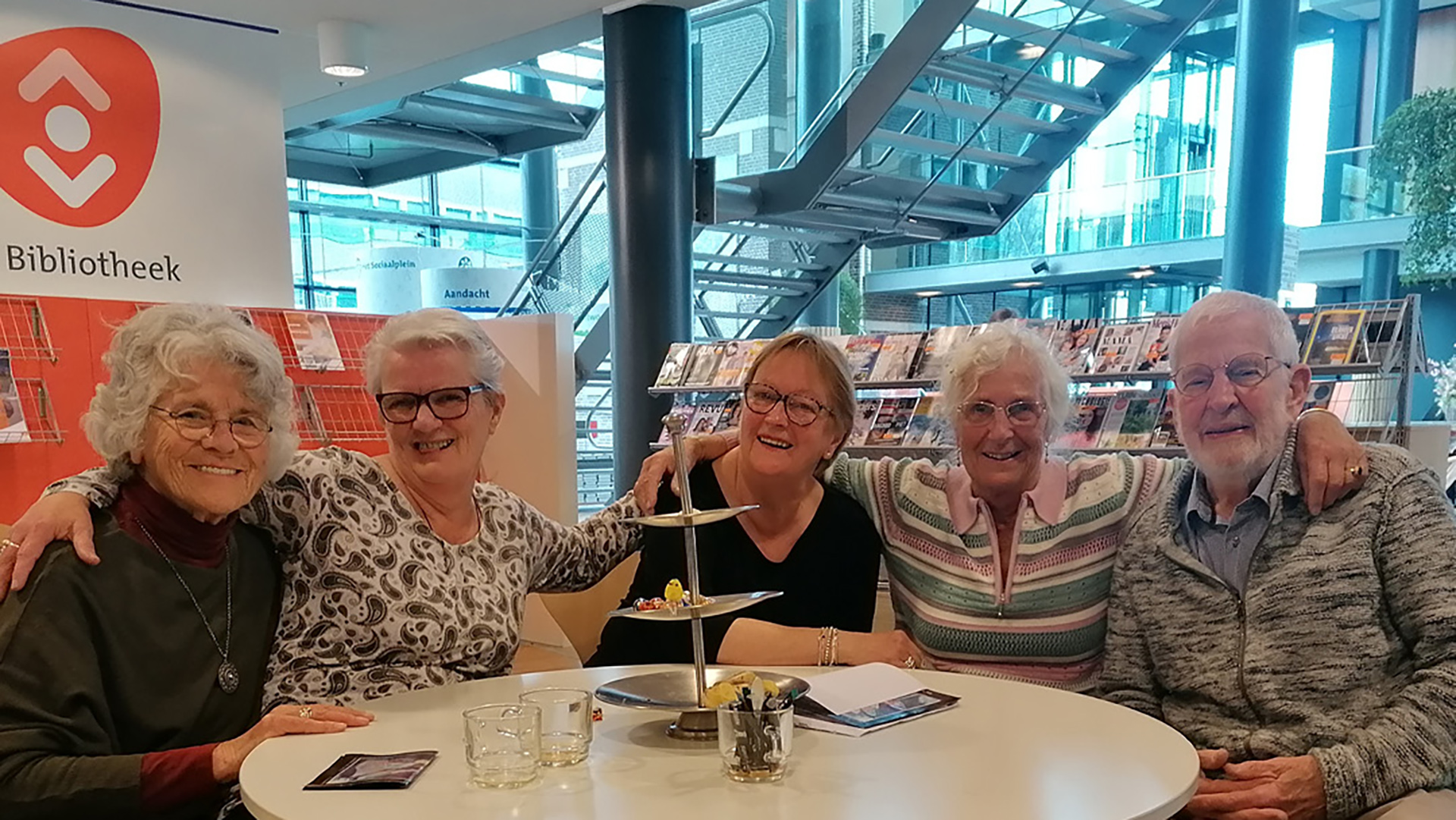 Vijf lachende ouderen zitten aan een tafel in een bibliotheek met een tijdschriftenrek op de achtergrond.