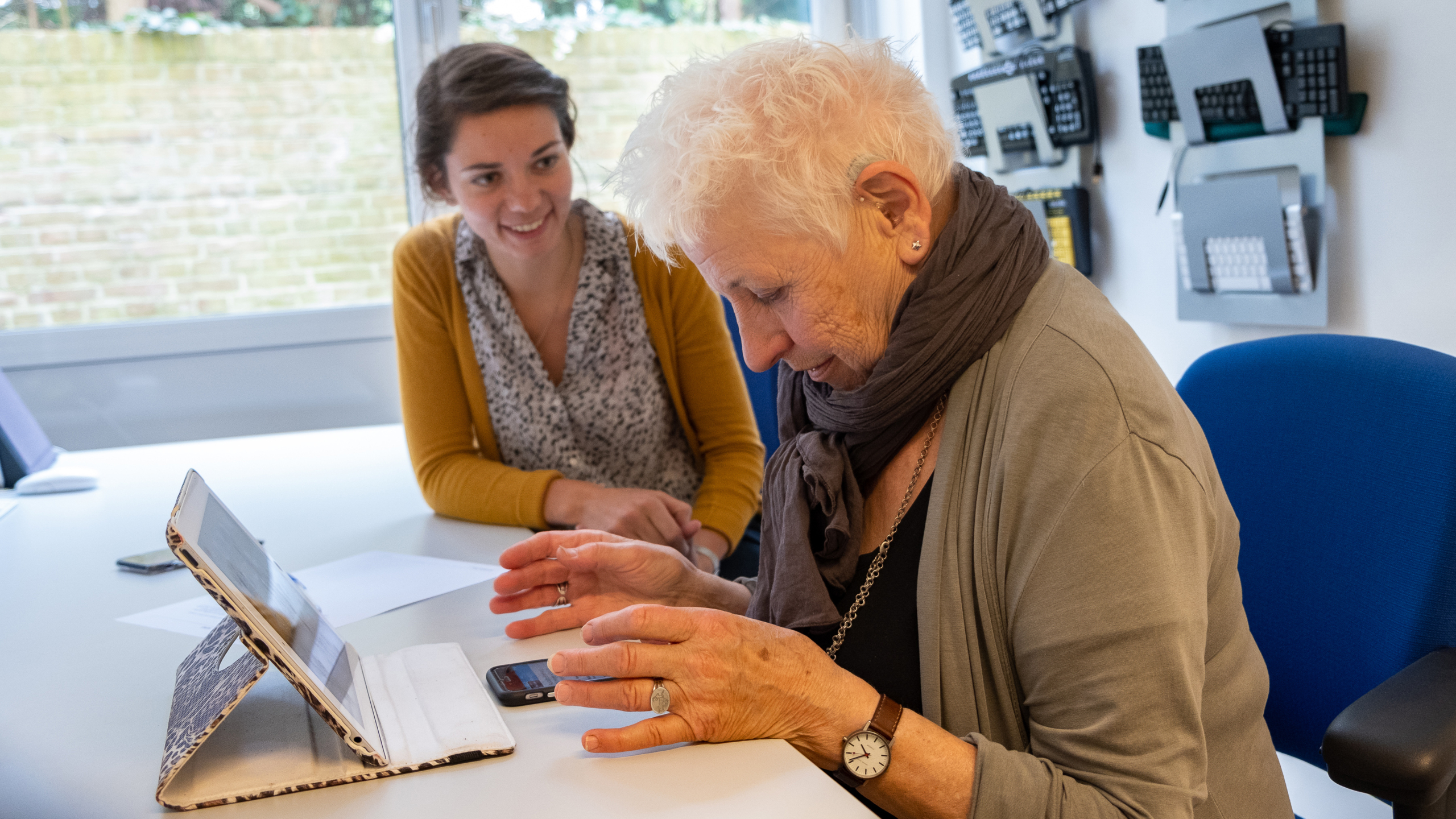 Een oudere vrouw gebruikt een tablet, terwijl een jongere vrouw naast haar zit en glimlacht.