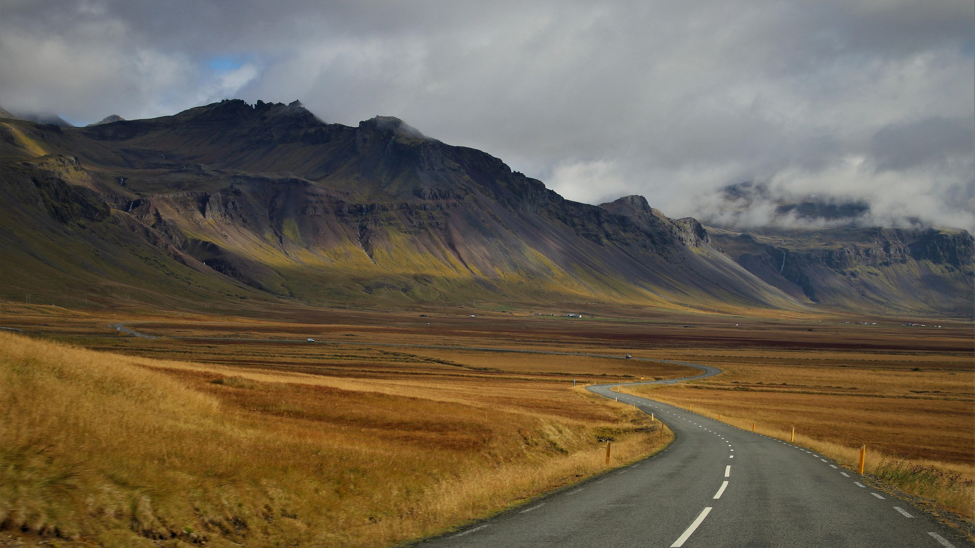 Kronkelende weg door een uitgestrekt landschap met bergen en lage bewolking op de achtergrond.