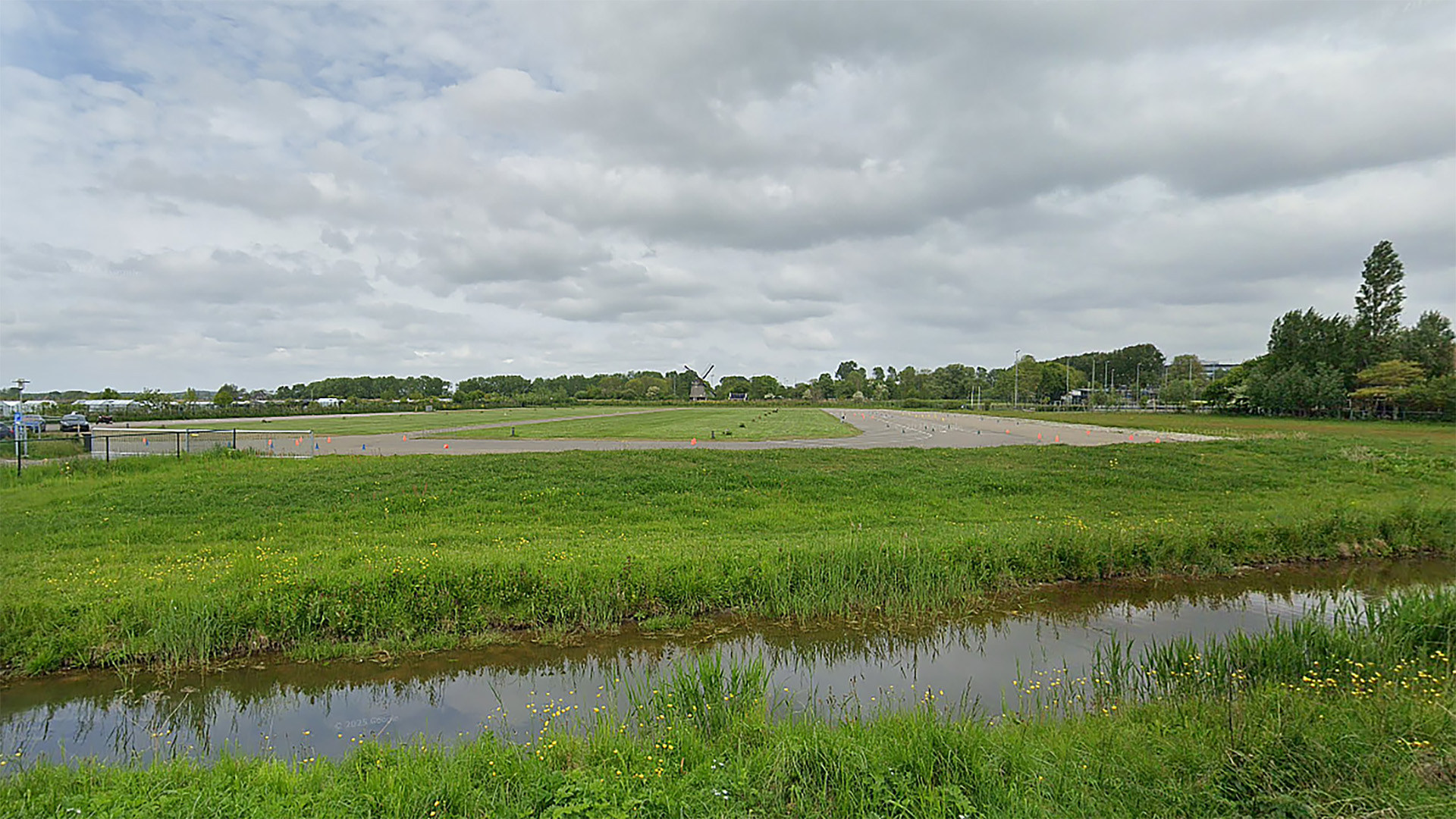 Een groen weiland met een sloot op de voorgrond en een lege parkeerplaats op de achtergrond onder een bewolkte hemel.