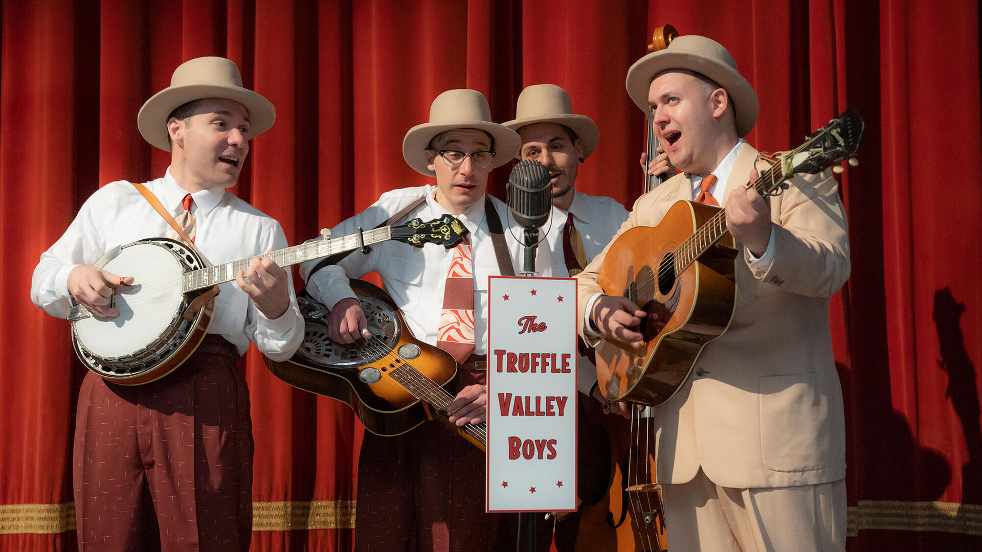 Vier muzikanten in vintage kleding spelen op banjo en gitaar voor een microfoon met het bord "The Truffle Valley Boys".