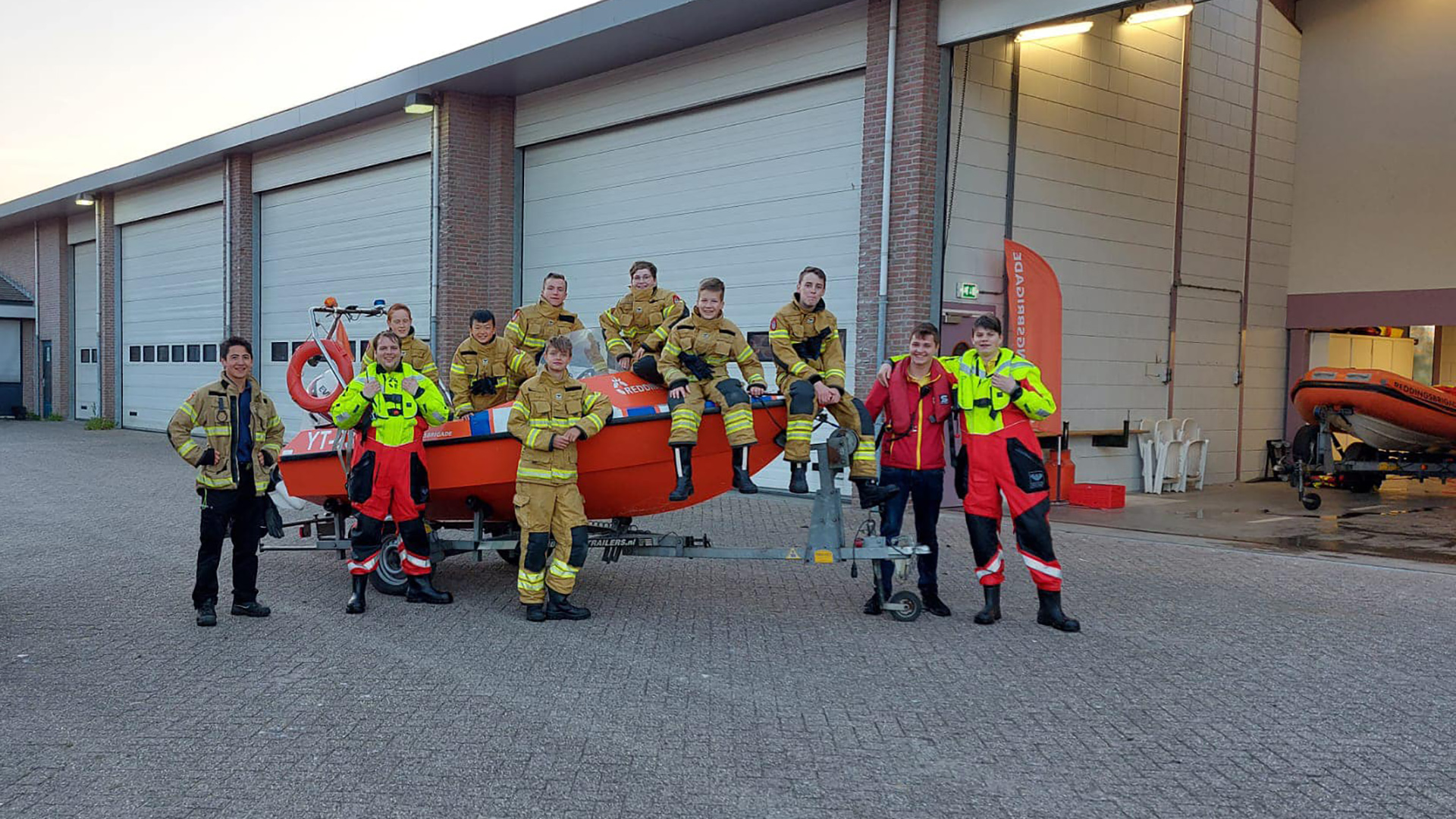 Brandweermensen poseren bij een oranje reddingsboot voor een garage met open deuren.