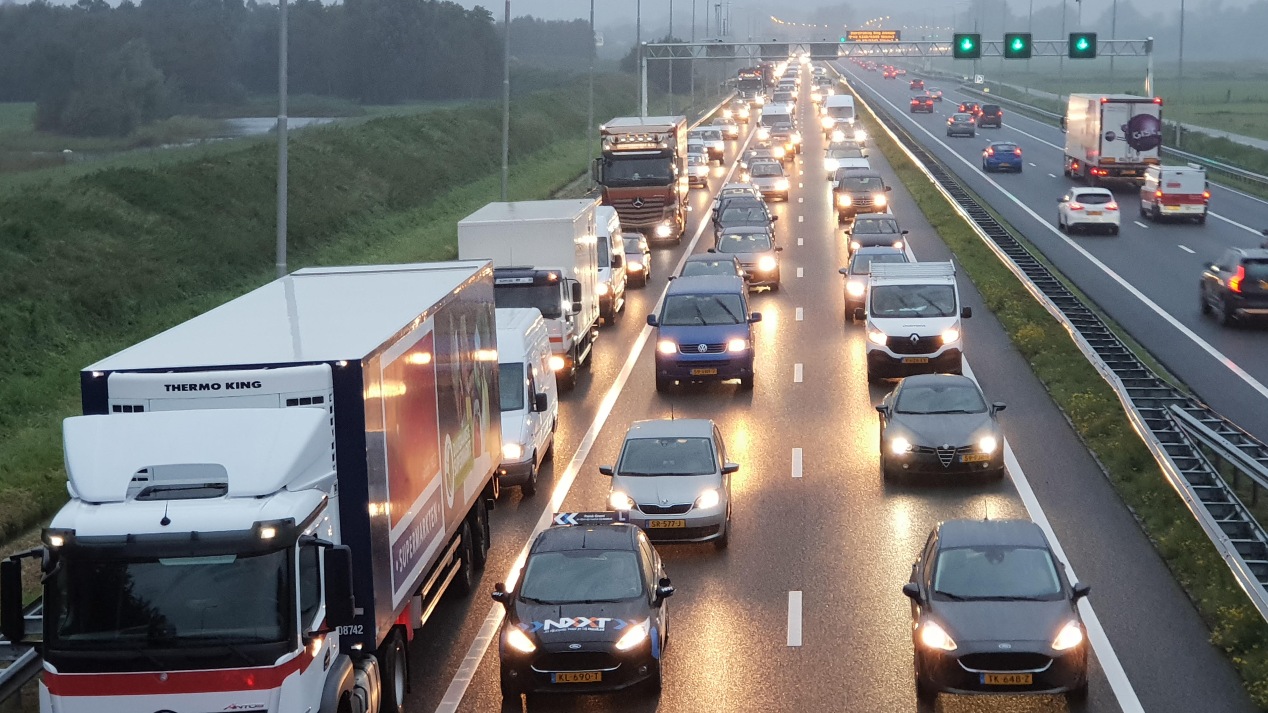 Drukke snelweg vol auto's en vrachtwagens in beide richtingen tijdens schemering, met groene verkeerslichten boven de rijbanen.