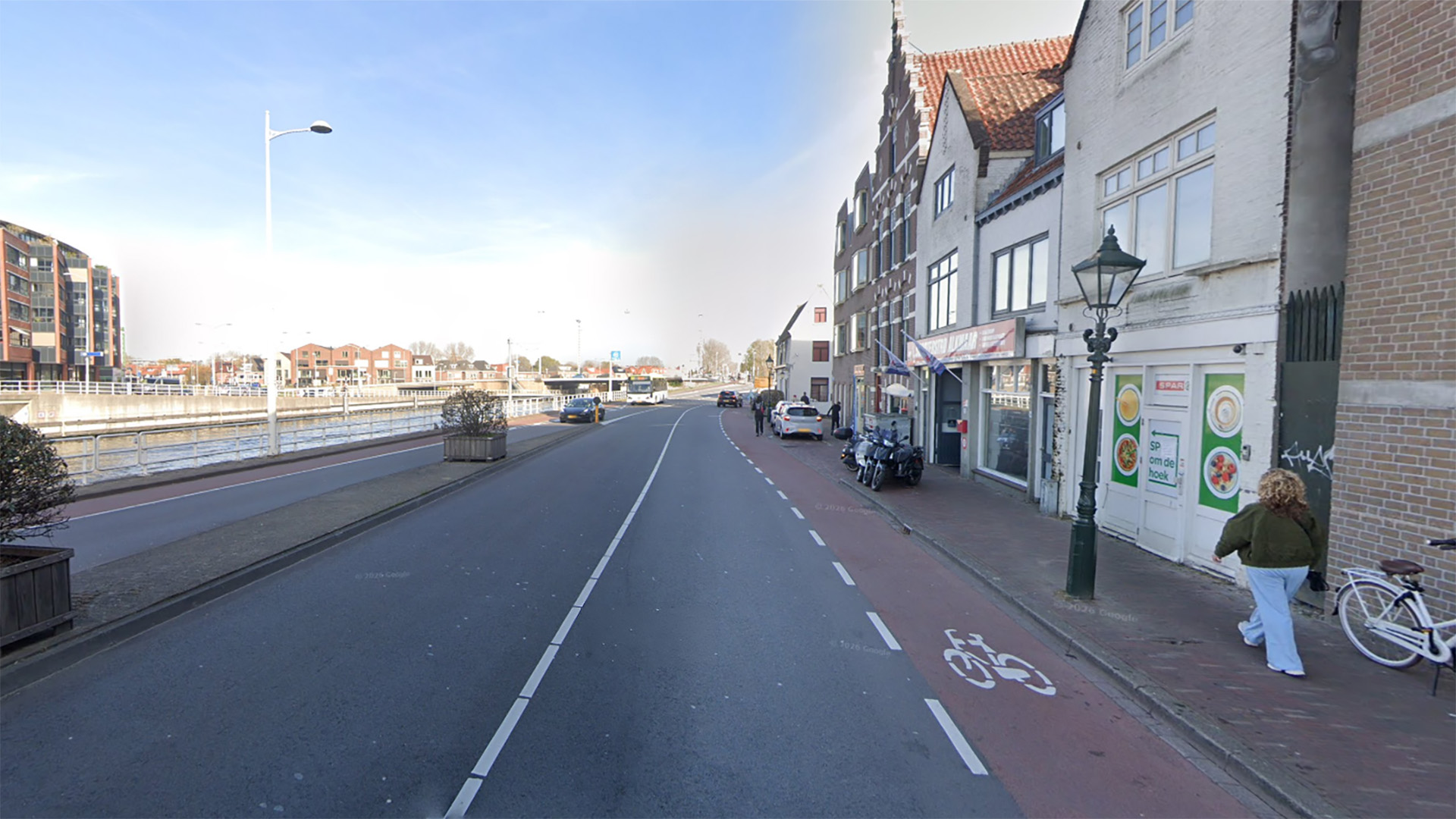 Een straatbeeld met een fietspad, een vrouw die loopt, winkels en een waterweg aan de linkerkant.