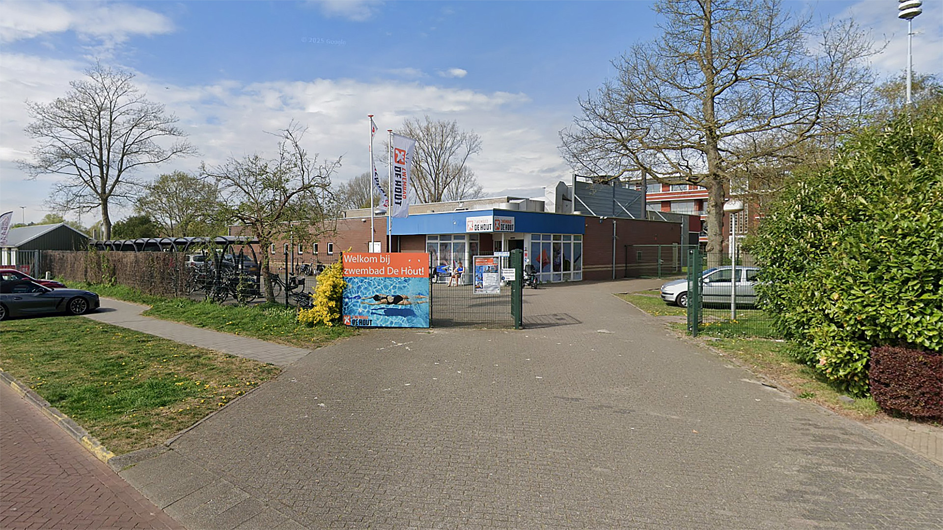 Entree van zwembad De Hout met een bord dat bezoekers verwelkomt, omgeven door fietsen en bomen.