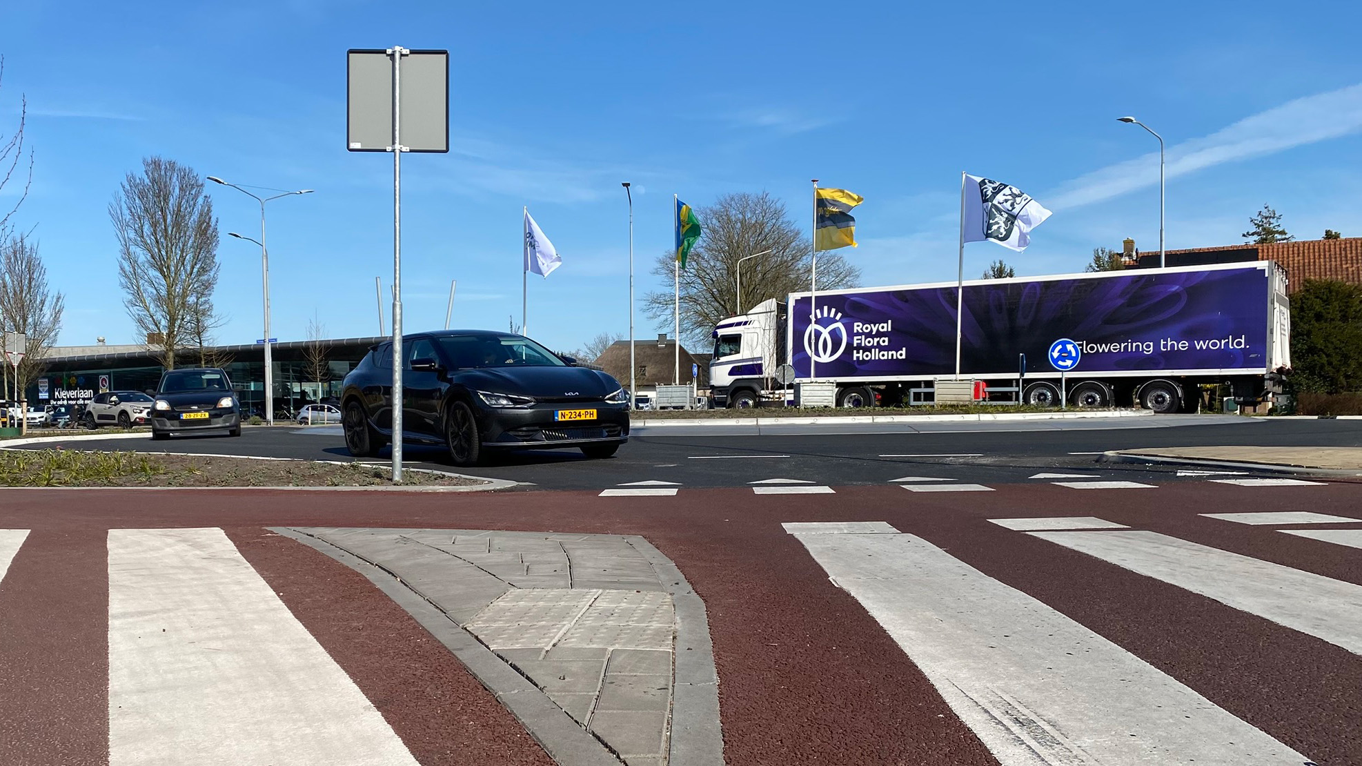 Auto bij rotonde met vrachtwagen van Royal Flora Holland op de achtergrond, zonnige dag met blauwe hemel.