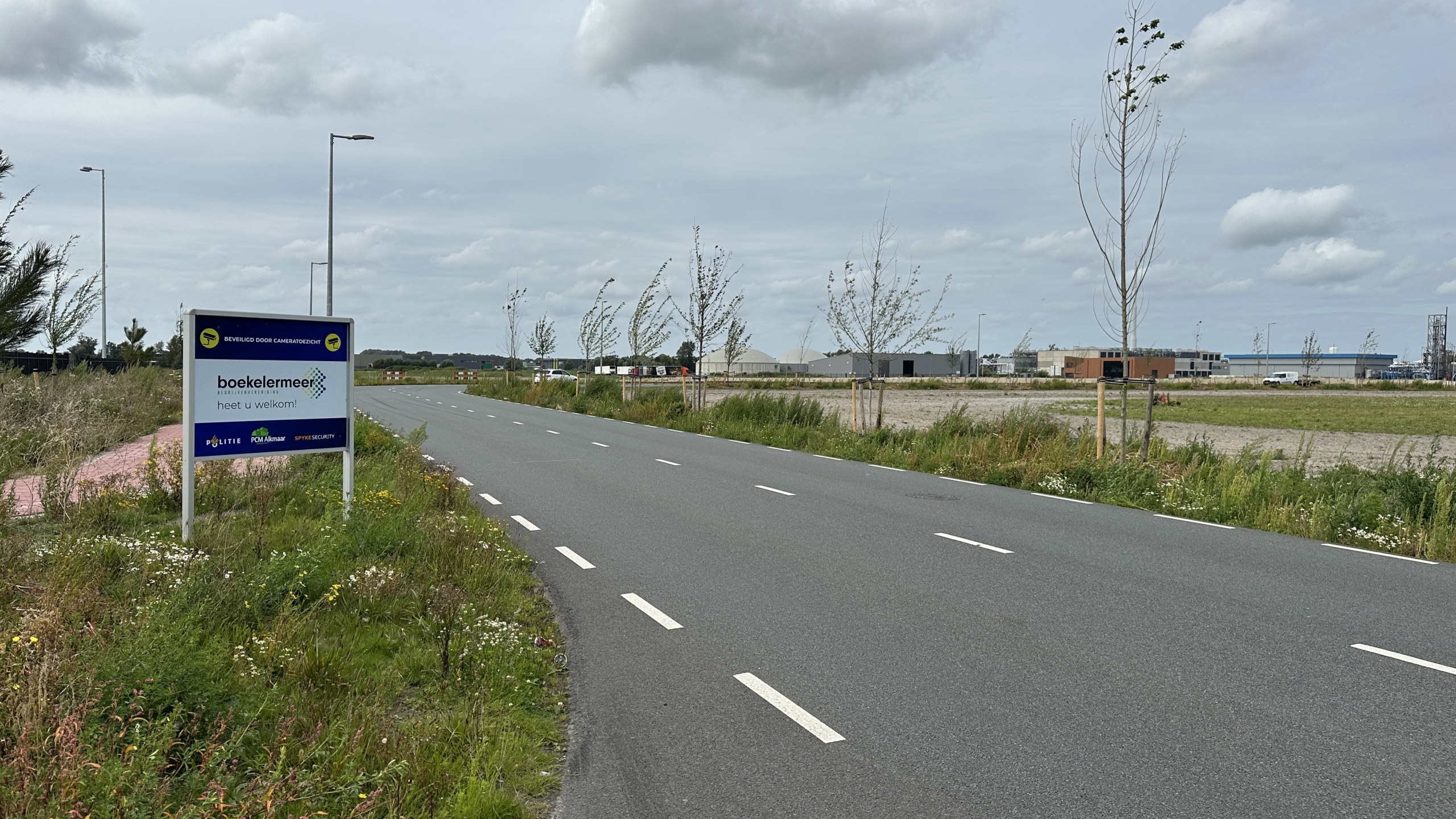 Bord langs een weg met de tekst "Boekelermeer heet u welkom!" in een industriële omgeving onder een bewolkte lucht.
