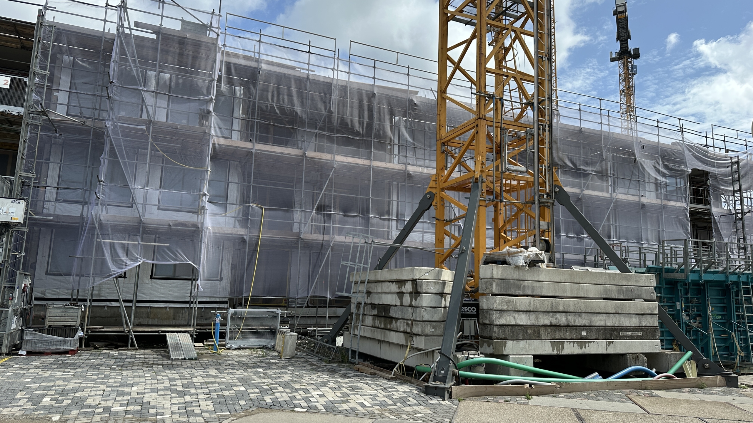 Bouwplaats met een hoog gebouw omgeven door steigers en netten, en een gele kraan op de voorgrond.